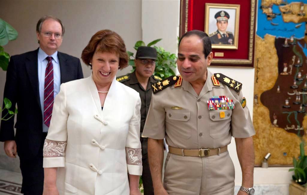 L'image montre une scène de rencontre entre plusieurs personnes dans un environnement officiel. Une femme souriante, vêtue d'une tenue claire, semble discuter avec un homme en uniforme militaire. Ce dernier porte des médailles et des insignias sur son uniforme. En arrière-plan, on peut voir un homme en tenue militaire additionnelle et une carte ou un tableau accroché au mur. L'ambiance semble amicale et détendue, suggérant un dialogue diplomatique ou militaire.