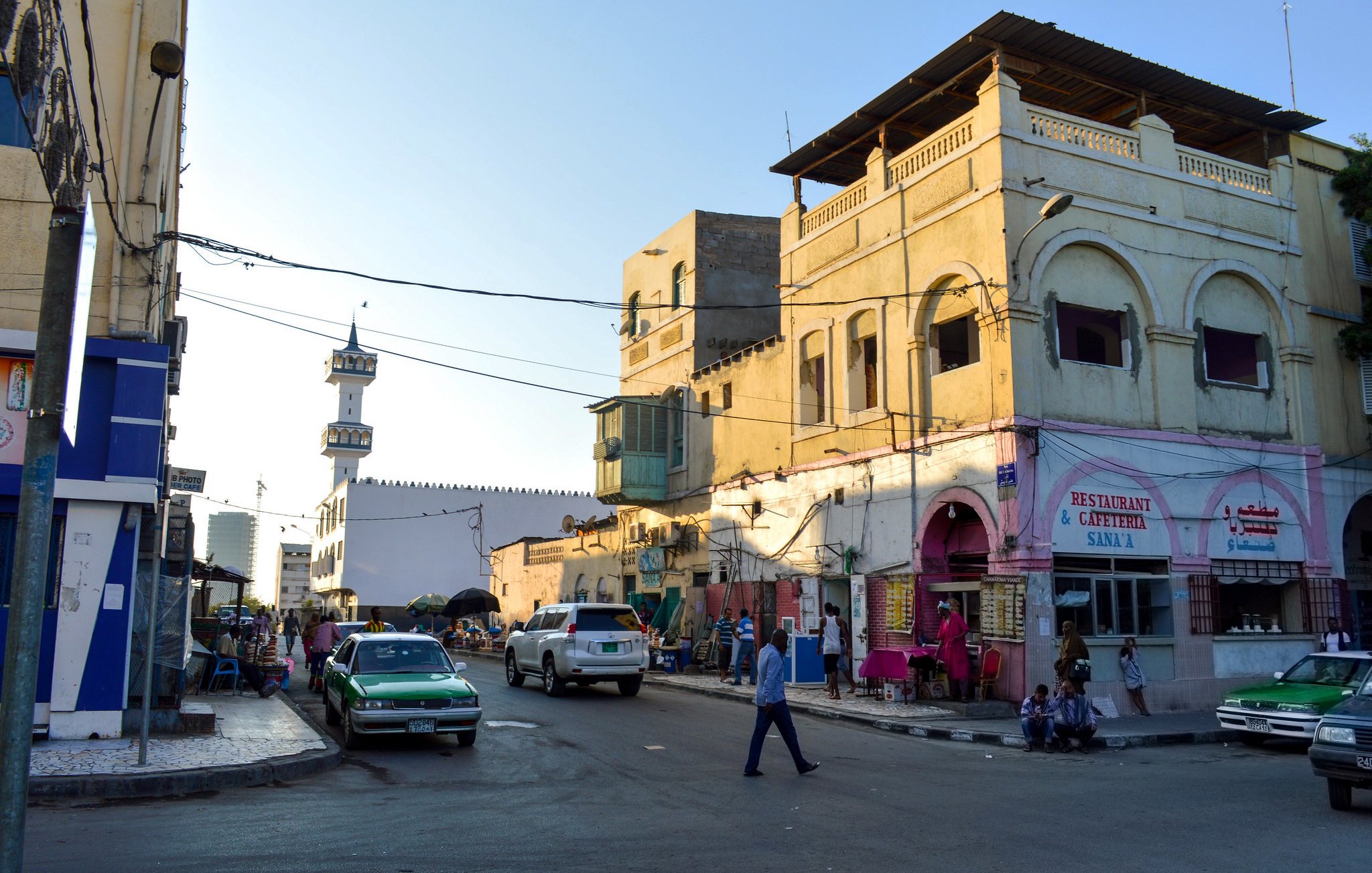 تظهر الصورة مشهداً لحياة الشارع في مدينة، حيث تتواجد المباني ذات الطراز المعماري التقليدي ذو الألوان الفاتحة. في الخلفية، يمكن رؤية مئذنة مسجد تُشير إلى وجود مكان للصلاة. في الشارع، تُظهر الحركة بعض المارة والسيارات، مما يعطي انطباعاً بالحيوية والنشاط. المناطق المحيطة بالأبنية تبدو مزدحمة، مع وجود عدد من المحلات التجارية والمقاهي. السماء زرقاء، مما يوحي بأن الوقت هو فترة الصباح أو بداية النهار.