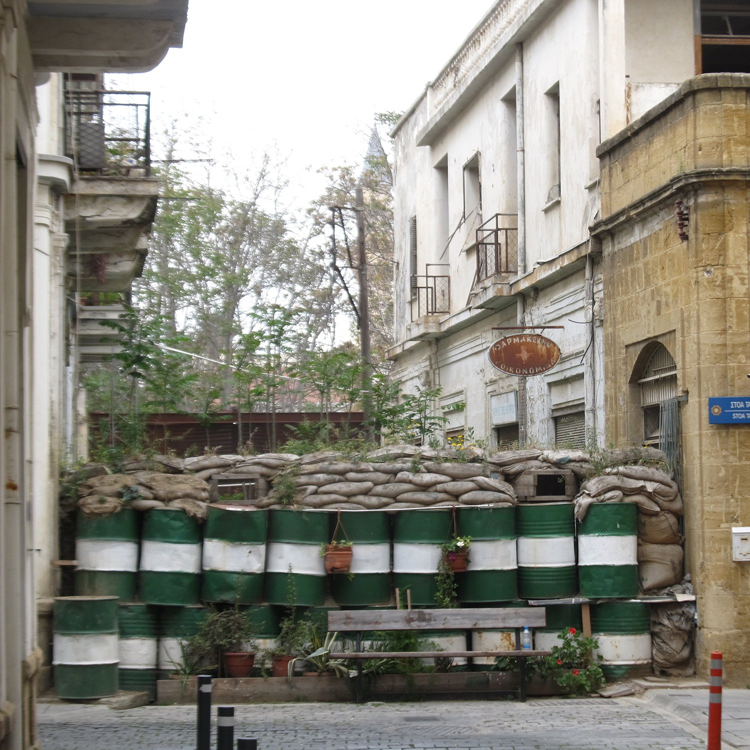 الصورة تظهر شارعاً ضيقاً في مدينة قديمة، حيث توجد مباني قديمة ذات تصميم معماري تقليدي. في نهاية الشارع، هناك حاجز مصنوع من أكياس الرمل وبراميل، مما يشير إلى وجود خطط للحماية أو الأمن. بعض النباتات تنمو حول الحاجز، مما يضفي لمسة من الحياة على المشهد. الألوان الرئيسية في الصورة هي درجات من البيج والأخضر، مما يعكس طابعاً قديماً وحالياً للمكان.