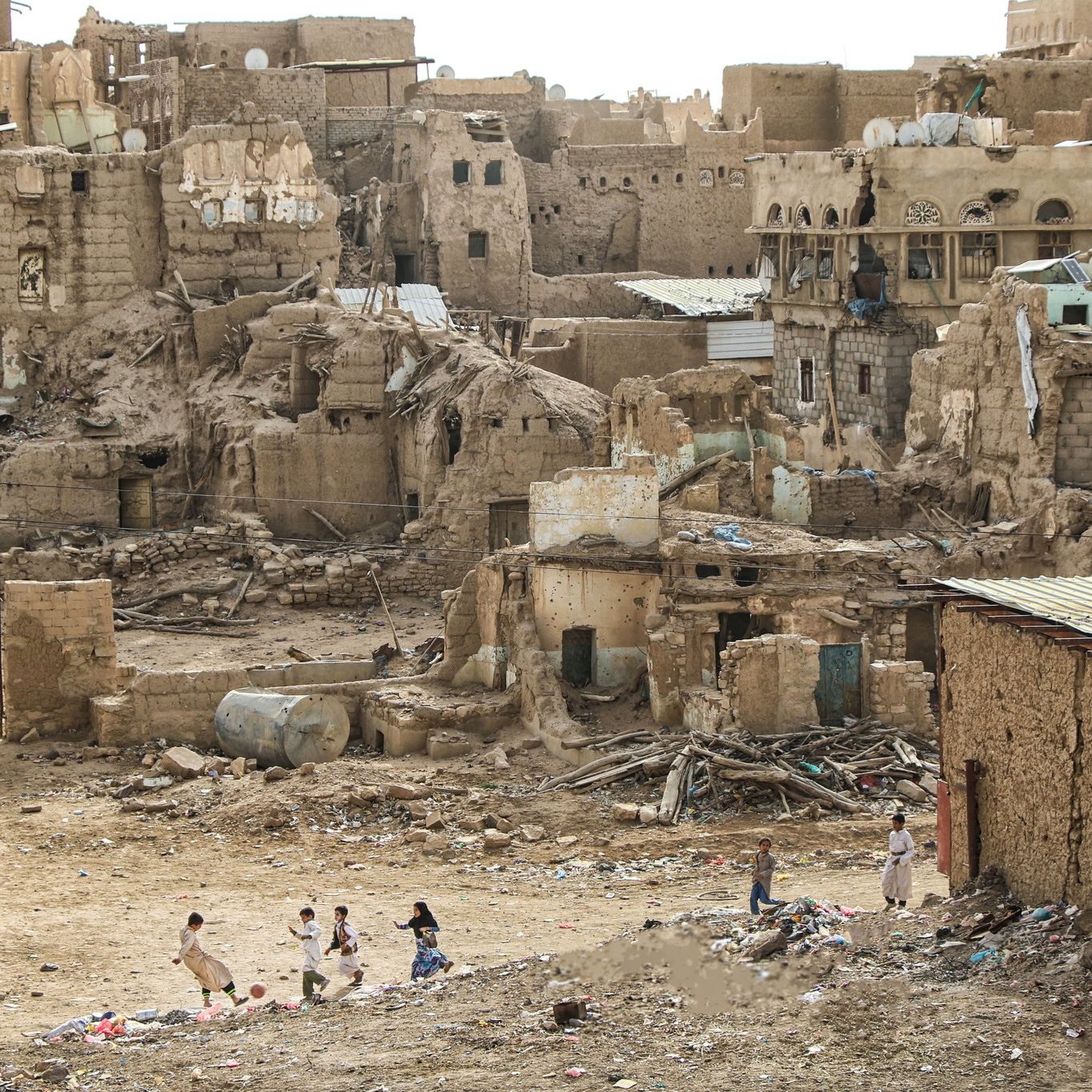 تصویر یک منطقه مخروبه و قدیمی را نشان می‌دهد که ساختمان‌ها و دیوارهای فرسوده و آجرهای شکسته در آن دیده می‌شود. در foreground، چند کودک در حال بازی هستند. زمین خشک و بی‌حاصل به نظر می‌رسد و بقایای ساخت و ساز و زباله‌ها در اطراف پراکنده شده‌اند. این منظر حس نابودی و تخریب را منتقل می‌کند و نمایی از زندگی در یک منطقه آسیب‌دیده را به نمایش می‌گذارد.