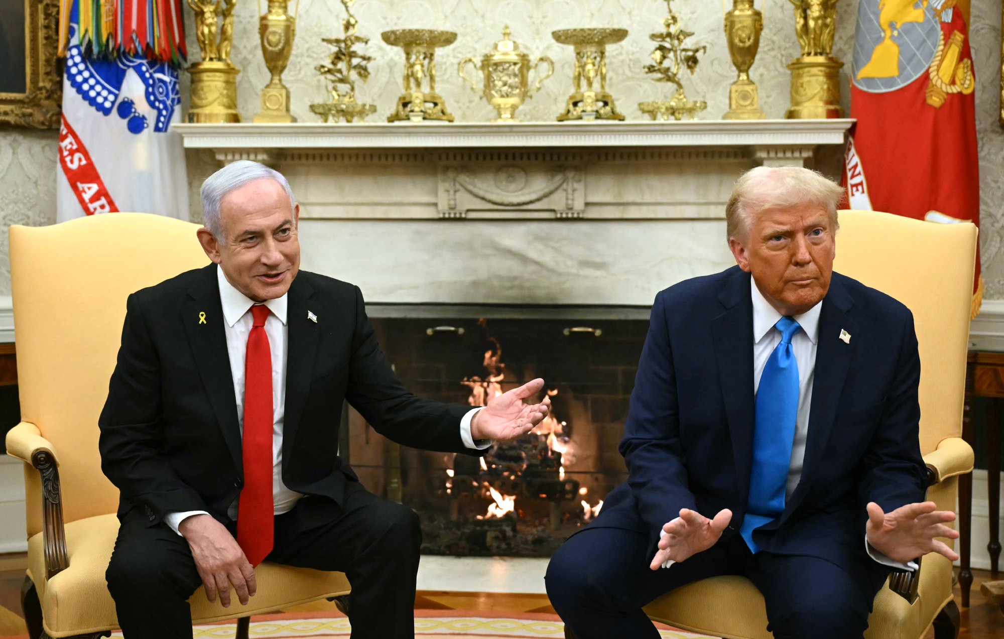 L'image montre deux hommes assis dans un bureau opulent, probablement dans le Bureau Ovale. L'un porte un costume noir avec une cravate rouge, tandis que l'autre est habillé en costume bleu avec une cravate rouge. Ils semblent engagés dans une conversation, avec des expressions sérieuses. En arrière-plan, on voit une cheminée décorée et divers objets sur des étagères. L'ambiance est formelle et politique.