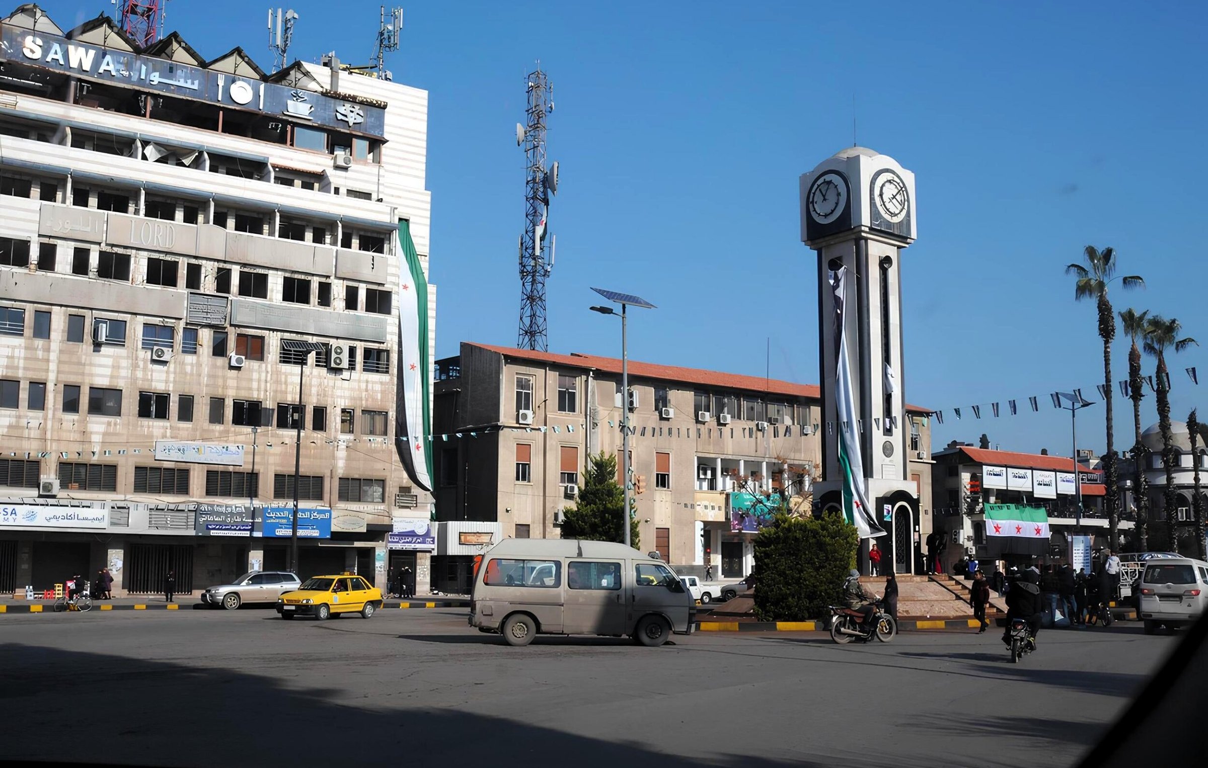 Una piazza con un grande orologio, edifici e alcuni veicoli in movimento.
