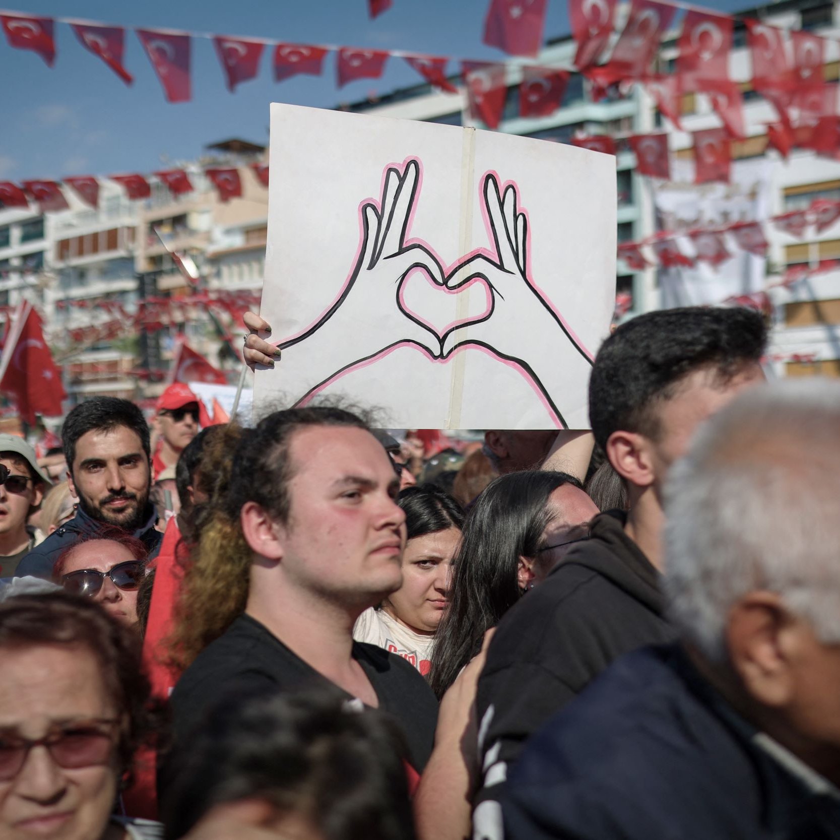 L'immagine mostra una grande folla riunita in un evento all'aperto, probabilmente una manifestazione o una celebrazione. Alcuni partecipanti sostengono bandiere turche, che sono di un rosso acceso, mentre un uomo in primo piano è visibile con una maglietta nera e guarda verso la telecamera. Dietro di lui, c'è un cartello che rappresenta due mani che formano un cuore, simbolo di amore o unità. L'atmosfera sembra essere festosa e piena di energia, con molte persone che condividono un momento di comunità.