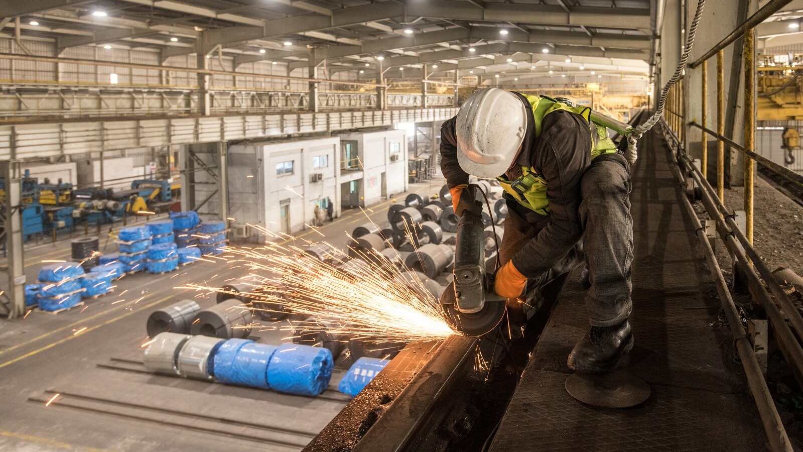 L'image montre un ouvrier en train de travailler dans un environnement industriel, probablement dans une usine. Il utilise une meuleuse, ce qui génère des étincelles. L'ouvrier porte un casque de sécurité et des gants, ce qui indique qu'il suit des mesures de sécurité adéquates. En arrière-plan, on peut voir des bobines de matériaux, ainsi que différents équipements et machines typiques d'un espace de travail industriel. L'éclairage de l'atelier semble naturel, avec des zones bien éclairées et d'autres plus sombres, mettant en valeur le côté dynamique et actif de l'environnement de travail.