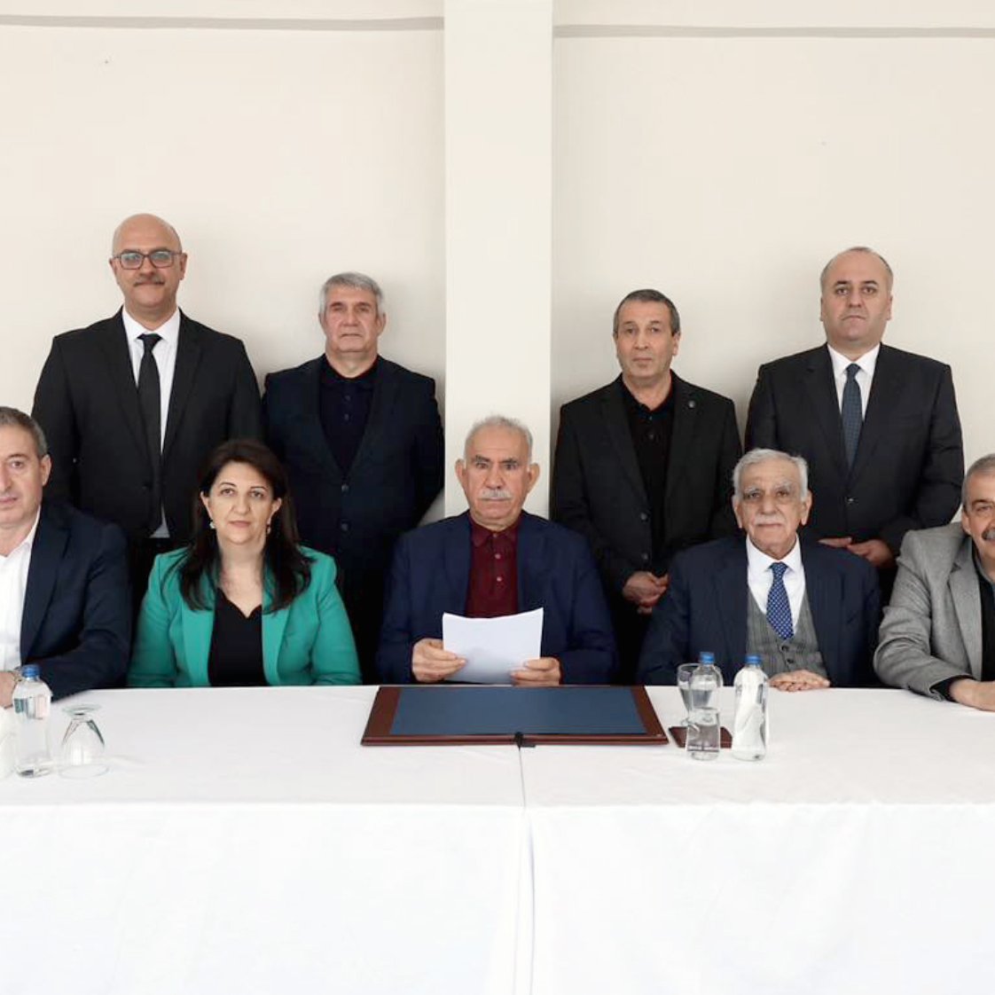 The image shows a group of twelve people seated around a table with a white cloth. They are dressed formally and appear to be engaged in a serious discussion or meeting.