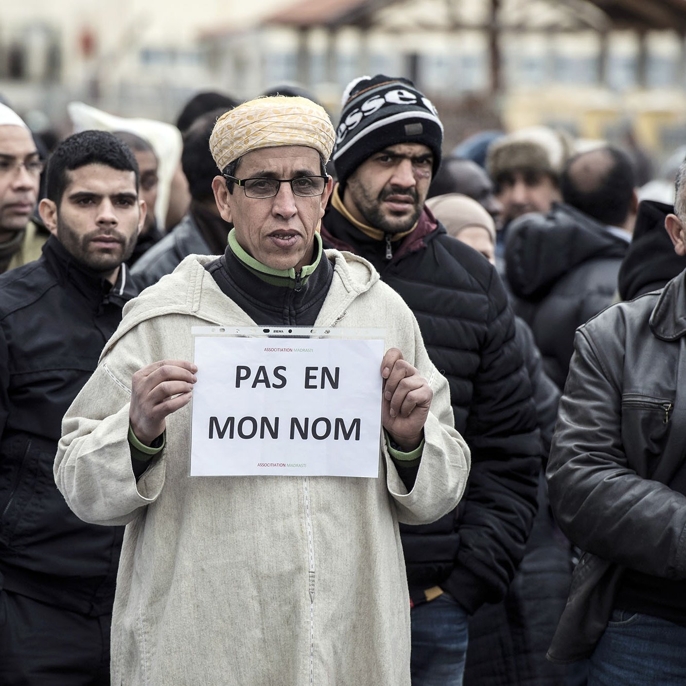 تظهر في الصورة مجموعة من الأشخاص يقفون في تجمع أو مظاهرة. في المقدمة، يوجد رجل يرتدي ملابس تقليدية ويقوم holding لافتة مكتوب عليها عبارة "PAS EN MON NOM" باللون الأبيض على خلفية بيضاء. يحيط به مجموعة من الرجال الذين يرتدون ملابس شتوية، ويبدو عليهم التوجه الجاد. تعكس الصورة أجواء من التعبير عن الرأي أو الاحتجاج.