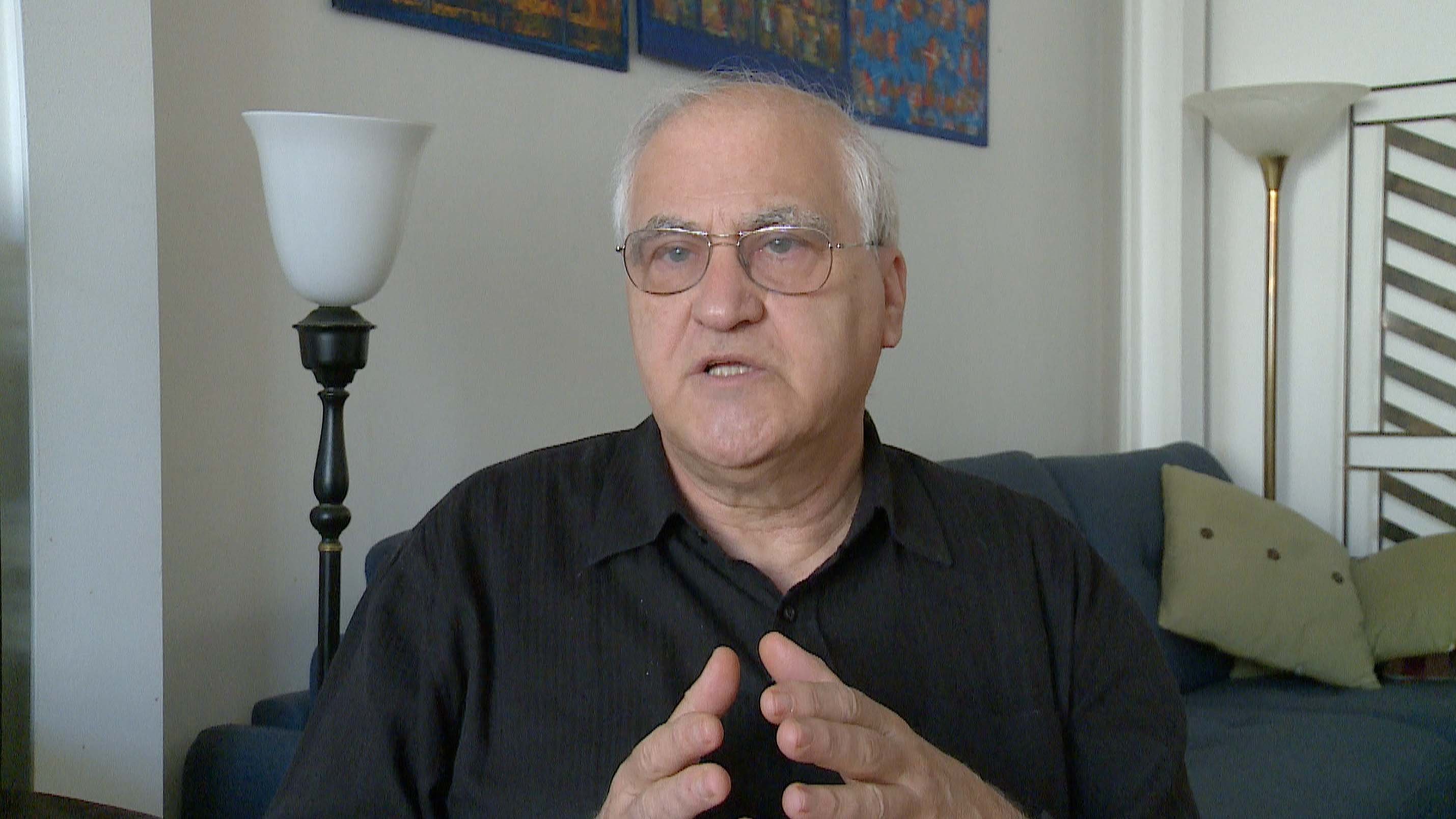 L'image montre un homme parlant avec des mains gesticulant, exprimant probablement une idée ou un point de vue. Il porte des lunettes et une chemise noire, et il semble attentif à la conversation. En arrière-plan, il y a un canapé bleu et une source de lumière, ce qui indique un environnement intérieur lumineux et accueillant. Des œuvres d'art sont accrochées au mur, ajoutant une touche décorative à la scène.