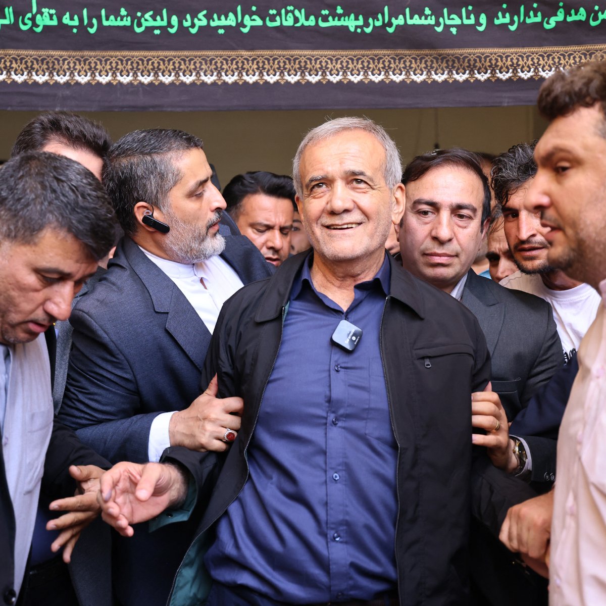 L'immagine mostra un gruppo di uomini in una situazione di affollamento. Al centro, un uomo di età avanzata sembra essere il focus dell'attenzione, mentre gli altri uomini lo circondano, alcuni di loro lo toccano o lo sostengono. L'atmosfera appare intensa e focalizzata, con uno sfondo decorato da un drappo o un banner che contiene testo in un'altra lingua. L'espressione dell'uomo centrale è di soddisfazione o di riconoscenza. 