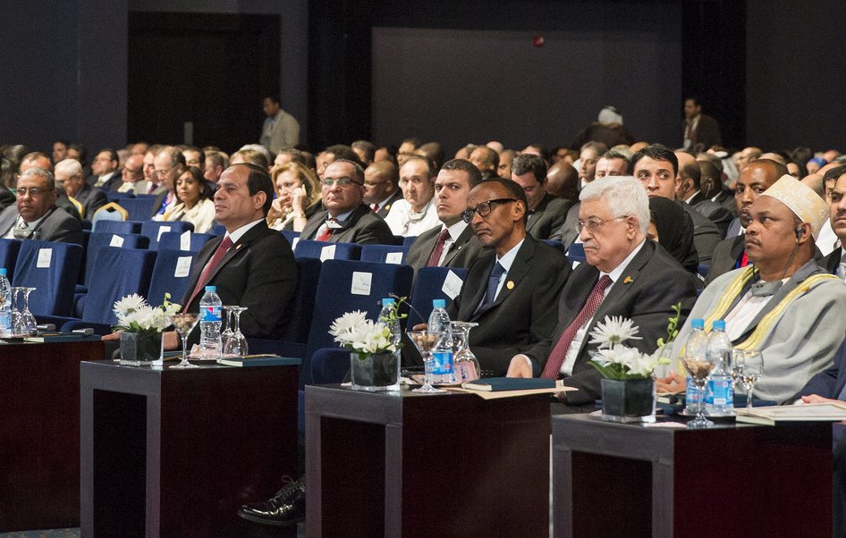 L'image montre une assemblée de personnes assises dans une salle de conférence. Les participants semblent attentifs et engagés, avec des tables devant eux, ornées de petites fleurs et de bouteilles d'eau. On peut voir plusieurs hommes en costume, ainsi que des femmes portant des vêtements formels. L'environnement est élégant, avec des chaises bleues et une disposition ordonnée. Les visages des participants expriment une concentration sur l'événement en cours.