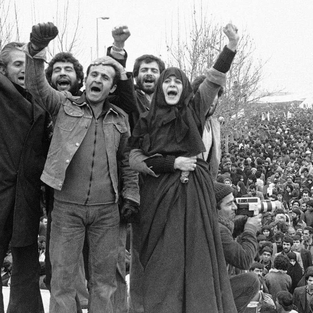 تصویر نمایی از یک تجمع بزرگ مردمی است که در میدان آزادی تهران برگزار شده است. در جلو، گروهی از افراد دست‌های خود را بالا برده و به نشانه اعتراض یا ابراز شادی فریاد می‌زنند. آن‌ها لباس‌های متفاوتی بر تن دارند و برخی از آن‌ها چهره‌های‌شان را خوشحال یا هیجان‌زده نشان می‌دهند. در پس‌زمینه، جمعیت زیادی دیده می‌شود که به سمت دوربین و افراد جلویی نگاه می‌کنند. بنای میدان آزادی نیز در پس‌زمینه قابل مشاهده است و جوّ کلی تصویر نشان‌دهنده شور و هیجان یا اعتراض اجتماعی است.