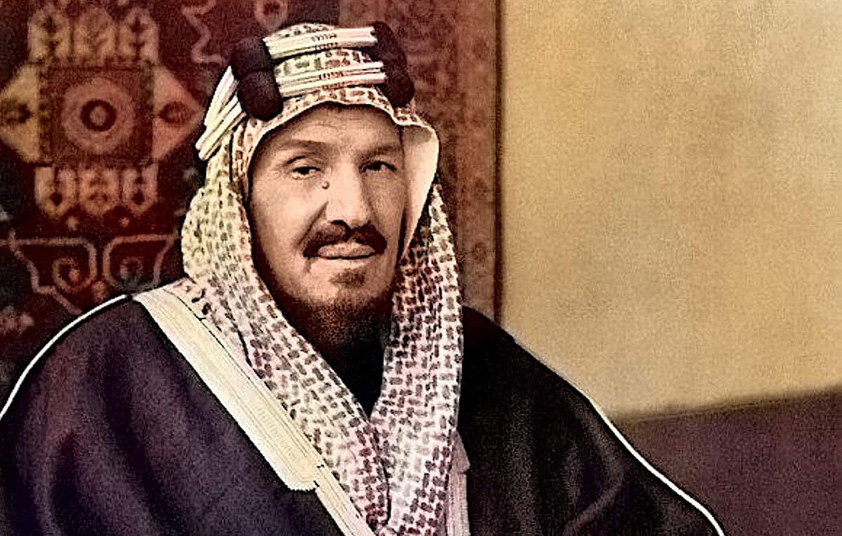 L'image montre un homme assis, portant un habit traditionnel. Il est vêtu d'une thobe sombre et d'un ghutrah, un couvre-chef caractéristique de la culture arabe. Il tient une canne et affiche une expression calme et posée. L'arrière-plan présente un motif décoratif qui renforce le contexte culturel de la scène. Les couleurs de l'image sont enrichies, donnant vie aux vêtements et à l'environnement.