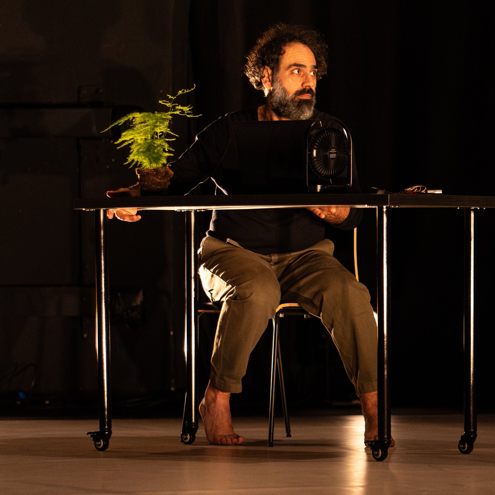 L'immagine mostra un uomo seduto a un tavolo in un ambiente di scena illuminato. Indossa una maglietta nera e pantaloni chiari, e sembra concentrato su un oggetto davanti a lui. Accanto a lui c'è una piccola pianta in vaso, mentre una ventola è posizionata sulla sua sinistra. L'illuminazione crea un'atmosfera drammatica, evidenziando sia l'uomo che gli oggetti sul tavolo. L'ambiente è scuro, con alcune luci che si riflettono sul pavimento.