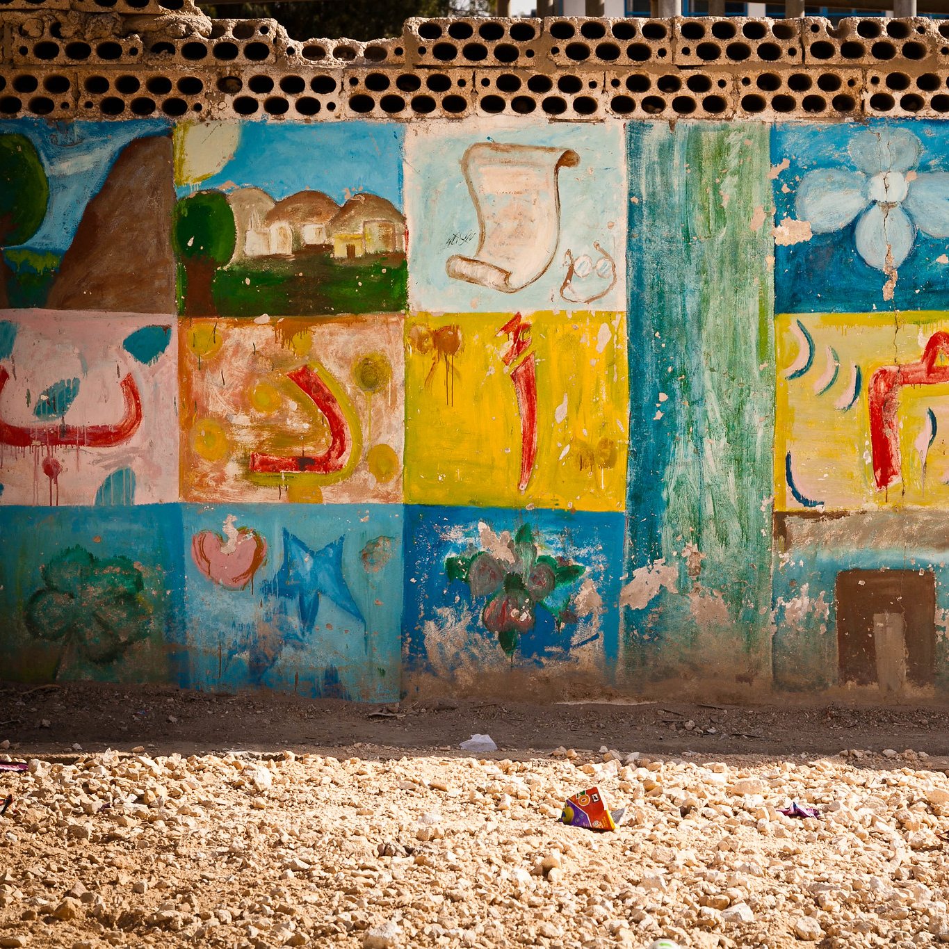 The image shows a wall decorated with colorful murals or paintings. The artwork includes various designs such as flowers, shapes, and possibly symbols or letters that are arranged in a grid pattern. The colors are vibrant, featuring shades of blue, green, yellow, and pink, giving the wall a lively and artistic feel. The surrounding area appears dusty or sandy, suggesting an outdoor environment. Overall, the scene conveys a sense of creativity and community.