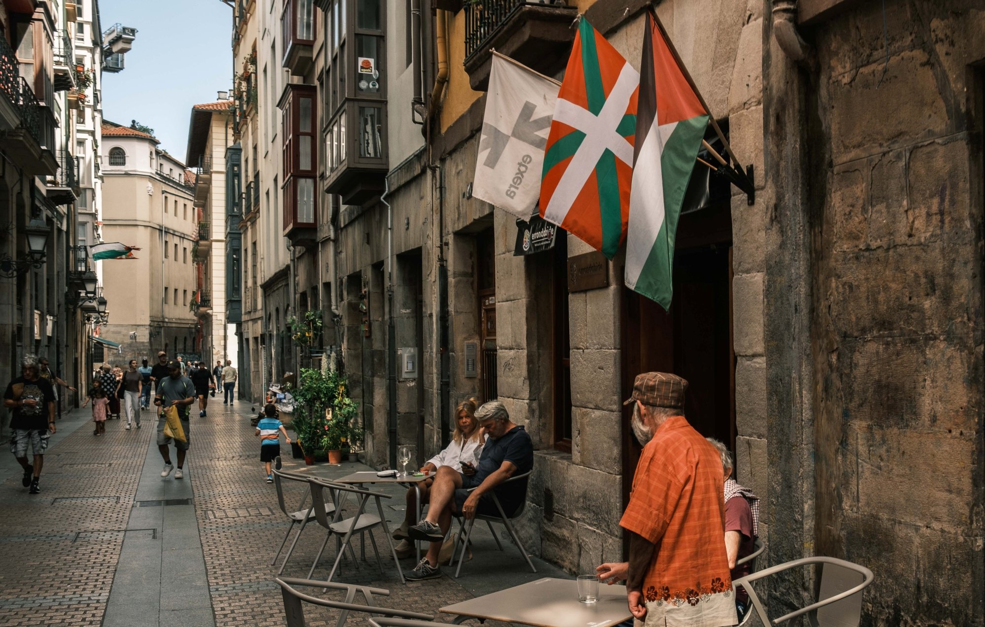 Calle estrecha con gente, mesas al aire libre y banderas ondeando en un ambiente urbano.