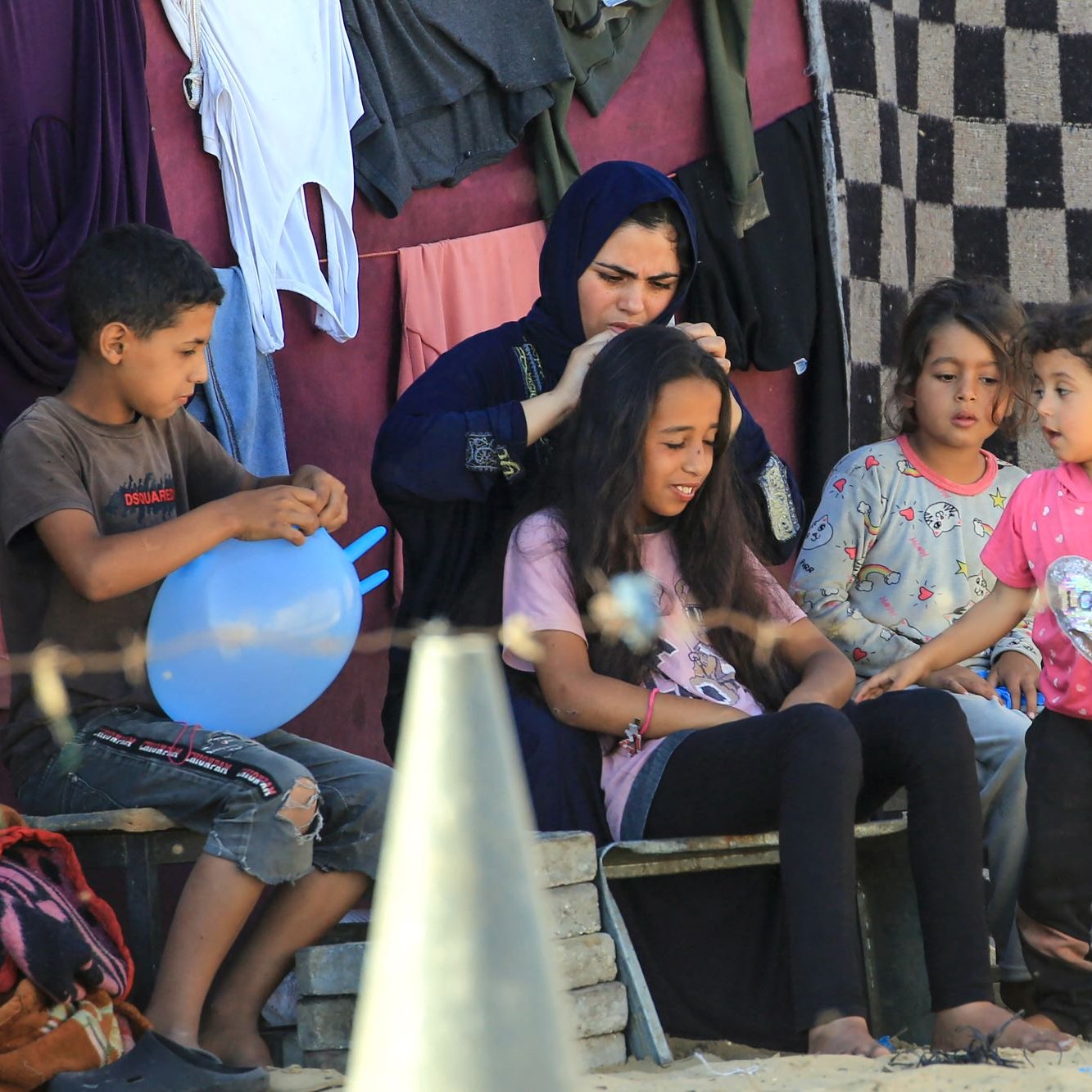 L'immagine mostra un gruppo di bambini e una ragazza seduti all'aperto, in un ambiente semplice. Una donna, vestita con un abito scuro, è intento a pettinare i capelli di un'altra ragazza, che sembra concentrata. Altri bambini sono presenti, alcuni giocano con un palloncino e altri osservano con interesse. Sullo sfondo, si possono vedere vestiti appesi e un ambiente rustico con pietre e teli. L'atmosfera sembra informale e familiare.