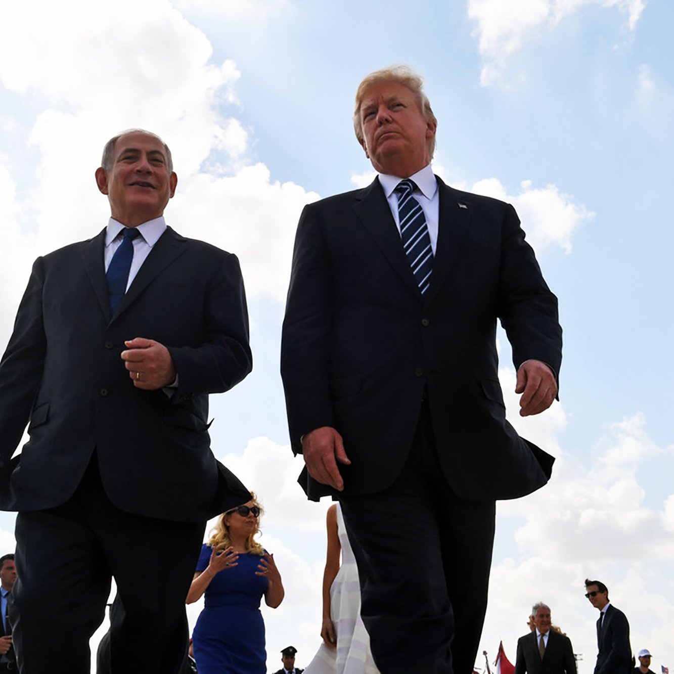 La imagen muestra a dos hombres caminando juntos al aire libre. Ambos visten trajes formales, con corbatas. Hay un grupo de personas detrás de ellos, y el cielo está parcialmente nublado. La escena transmite una sensación de formalidad y camaradería.