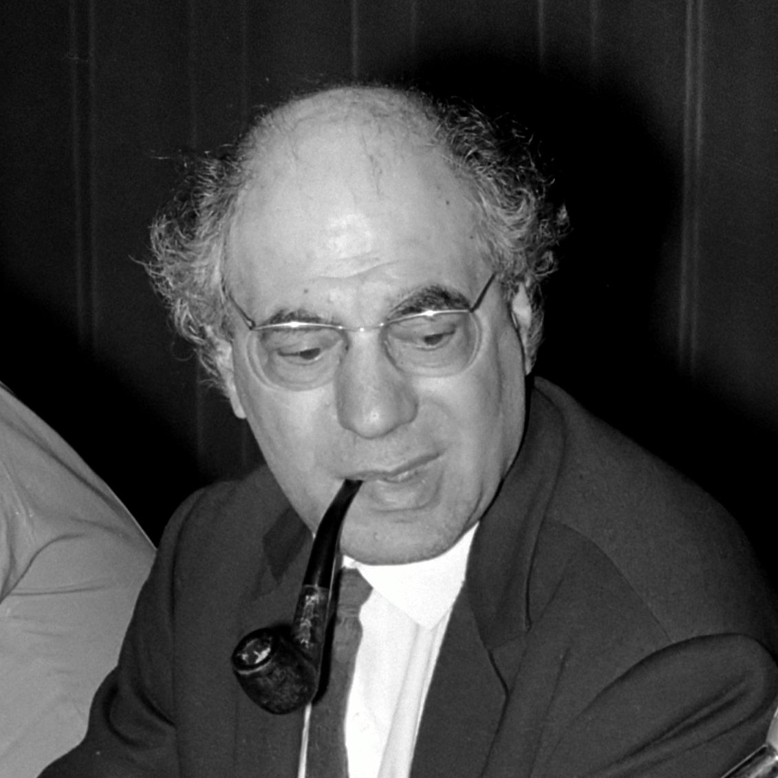 تظهر في الصورة رجل يجلس على طاولة، يبدو أنه في وضعية نقاش أو اجتماع. لديه شعر رمادي ومجعد، ويرتدي نظارات ويشعل غليونًا. يرتدي بدلة داكنة مع قميص وروابط. تعكس تعابير وجهه تركزًا واهتمامًا. البيئة المحيطة تبدو بسيطة، مع تفاصيل غير واضحة في الخلفية.