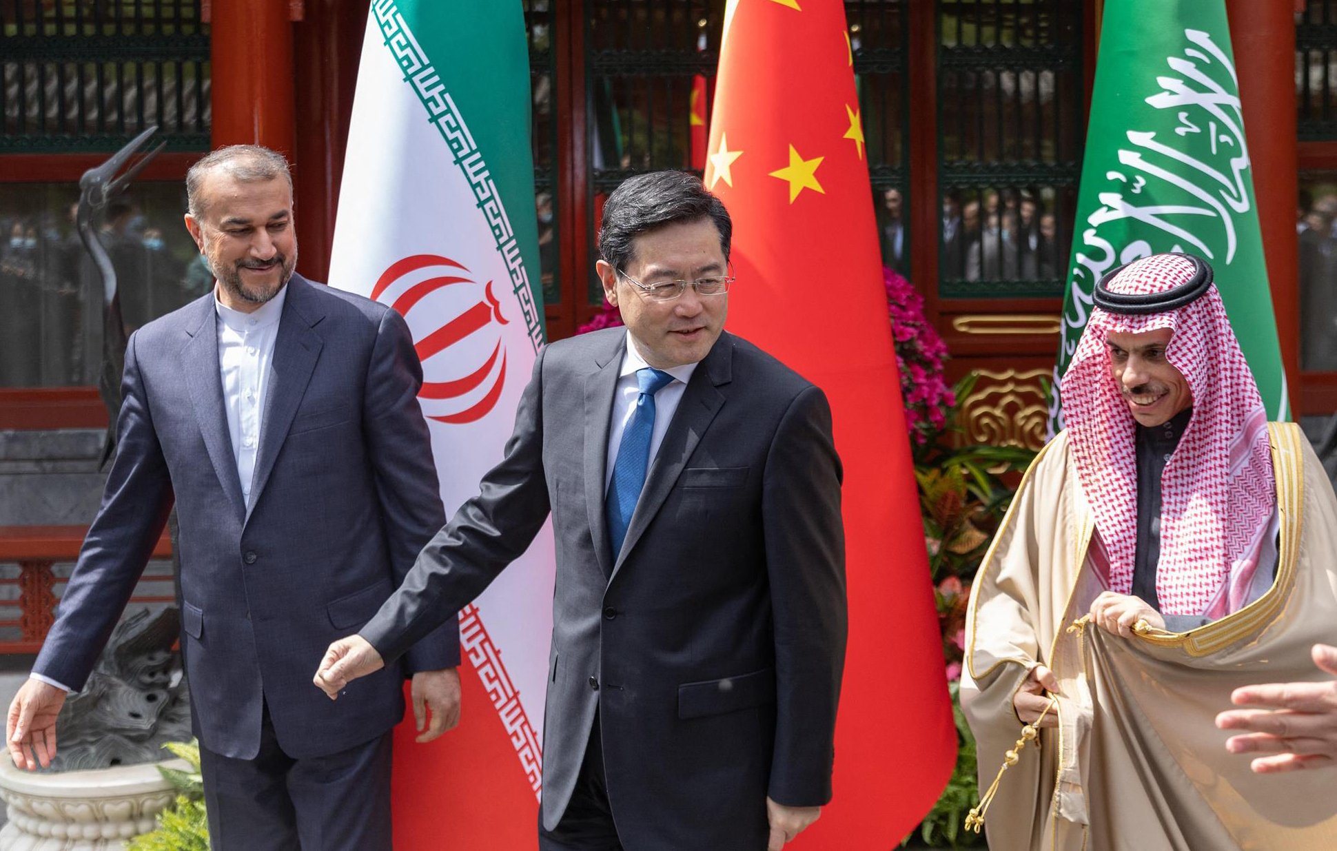 L'image montre trois hommes en costume, se tenant côte à côte devant des drapeaux. On devine que ces hommes représentent respectivement l'Iran, la Chine et l'Arabie Saoudite, compte tenu des drapeaux en arrière-plan. Ils semblent être en discussion lors d'un événement officiel. La scène est colorée, avec des éléments décoratifs comprenant des fleurs. Les hommes affichent des expressions sérieuses et engagées.