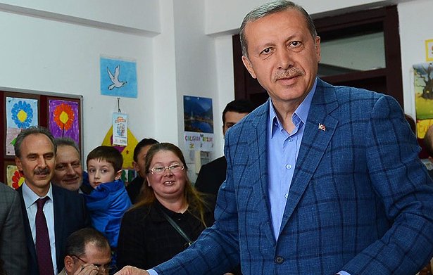 L'image montre un homme d'âge moyen portant un costume bleu. Il est debout, tenant un bulletin dans sa main, et semble être dans une salle de vote ou un endroit similaire. Il y a plusieurs personnes en arrière-plan, dont une femme avec des lunettes, un enfant et d'autres adultes, qui regardent l'homme avec intérêt. L'environnement est chaleureux et coloré, avec des affiches accrochées aux murs.