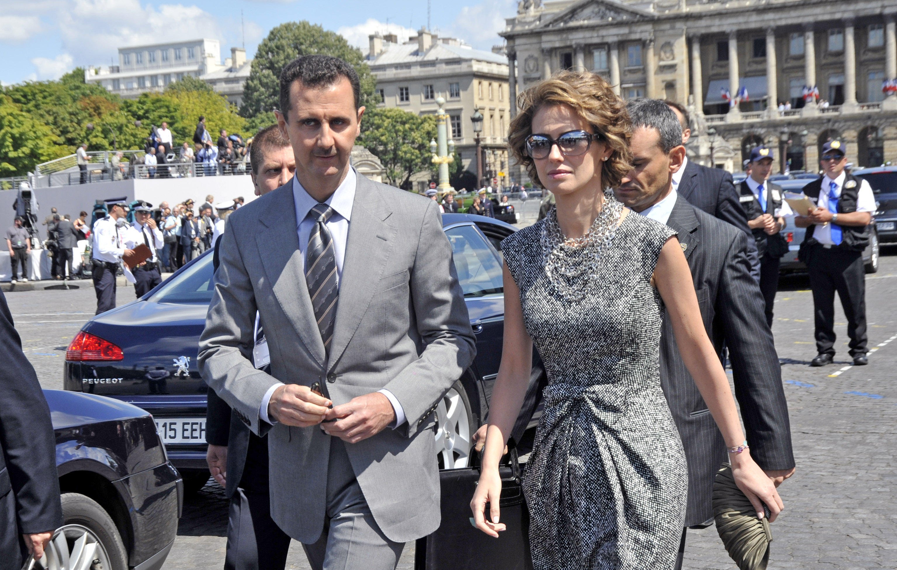 The image shows a man and a woman walking together in an outdoor setting. The man is dressed in a grey suit and tie, while the woman is wearing a stylish, patterned dress, sunglasses, and holds a handbag. They appear to be in a public area, possibly during a formal event, with a group of people in the background, including security personnel. The architecture behind them suggests a significant building or landmark.