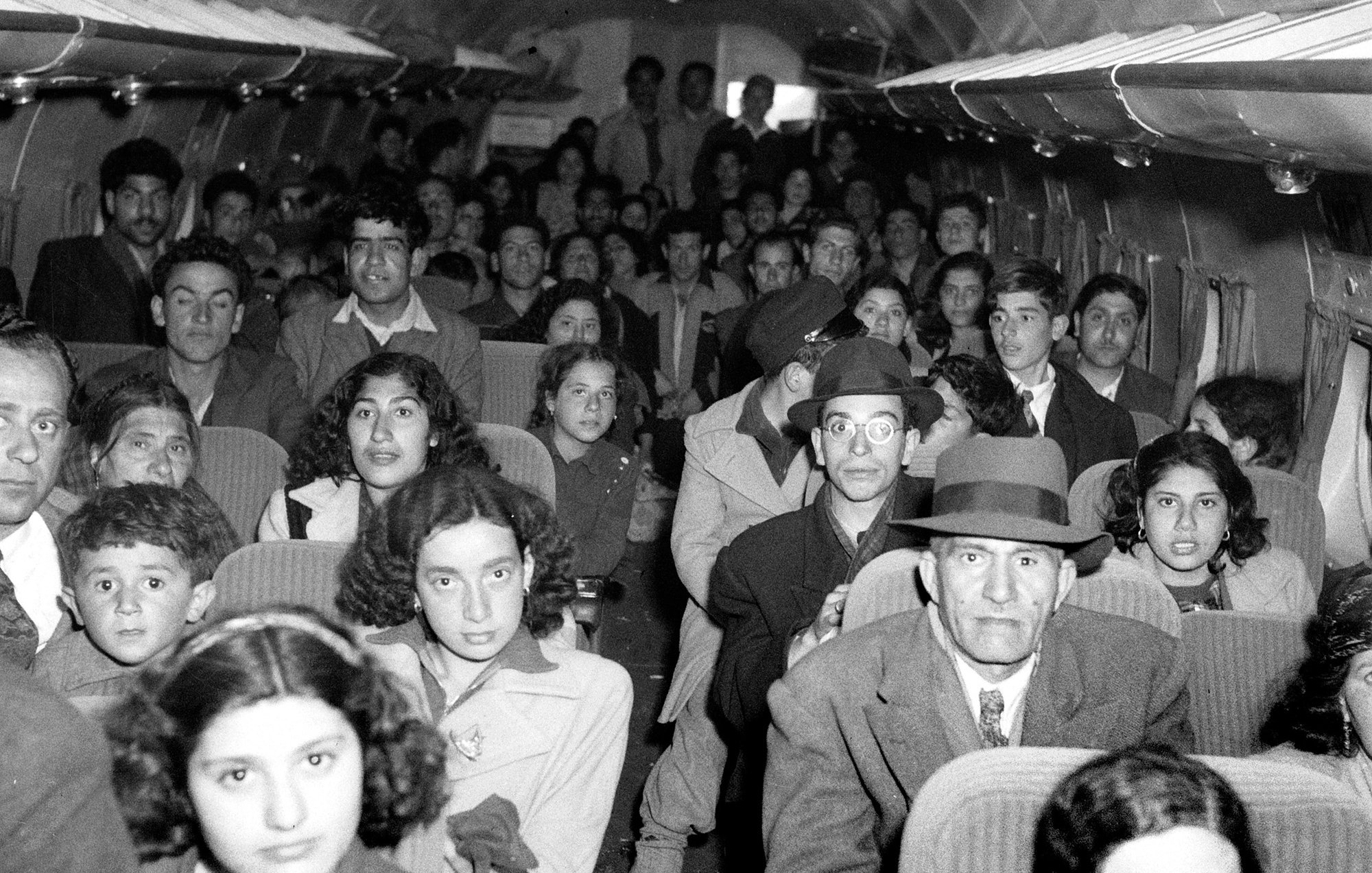 الصورة تُظهر مجموعة كبيرة من الأشخاص داخل طائرة، حيث يبدو أن معظمهم يجلسون في مقاعدهم. هناك تنوع في الأعمار، بدءًا من الأطفال إلى البالغين. يمكن رؤية تعبيرات مختلفة على وجوه الركاب، حيث يبدون جميعًا في حالة انتظار أو استعداد للسفر. تصميم الطائرة بسيط، مع نوافذ صغيرة فوق مقاعد الركاب. الأزياء تبدو تقليدية، مما يعطي انطباعًا عن زمن قديم.