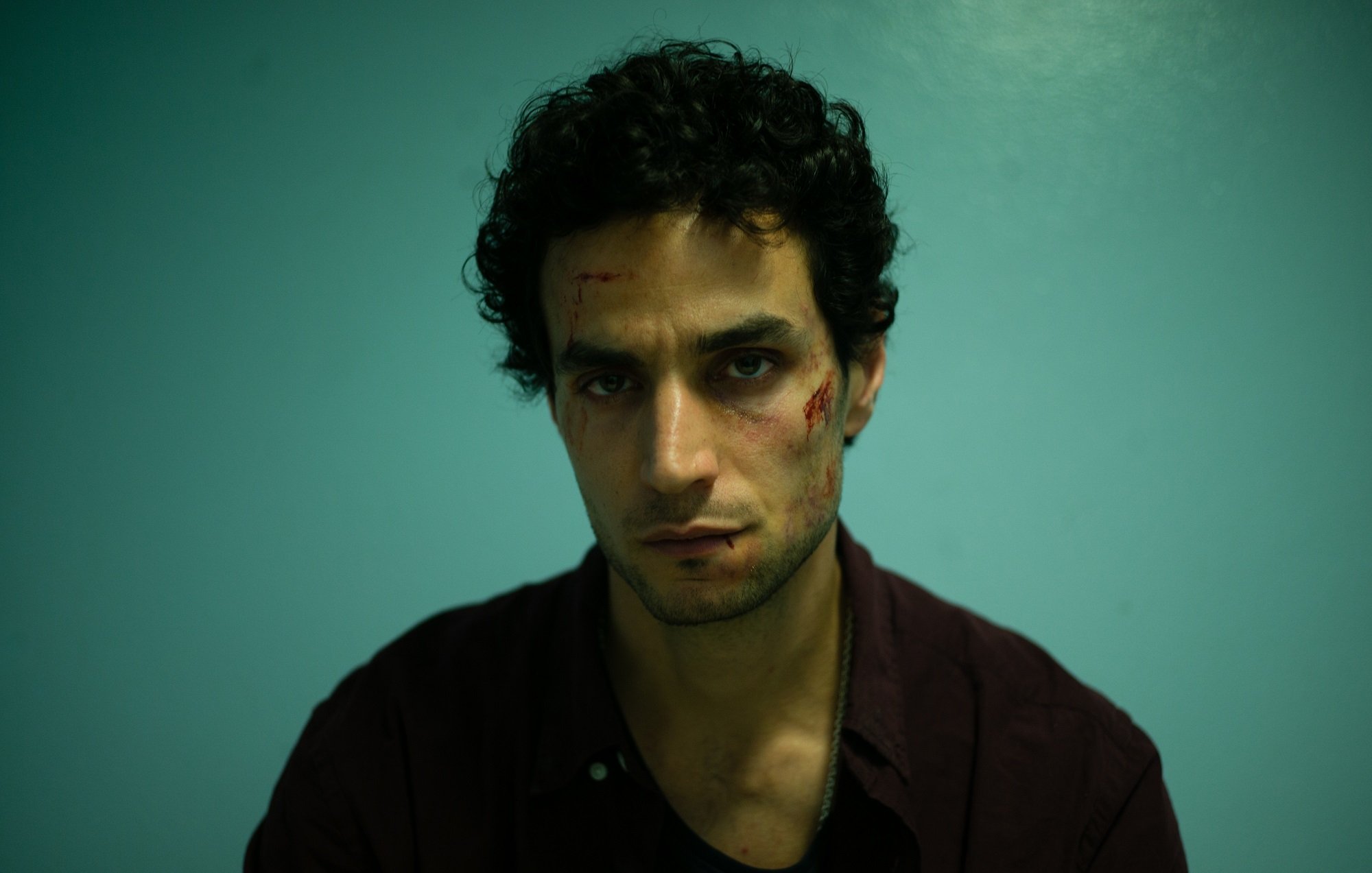 L'image montre un homme assis devant un mur de couleur bleu clair. Il a des cheveux noirs et bouclés, et son expression faciale semble intense ou préoccupée. Son visage présente des éraflures ou des blessures visibles, ce qui suggère qu'il a traversé une situation difficile. Il porte une chemise foncée et a l'air fatigué ou en détresse. L'éclairage est dramatique, mettant en avant ses traits et les blessures sur son visage.
