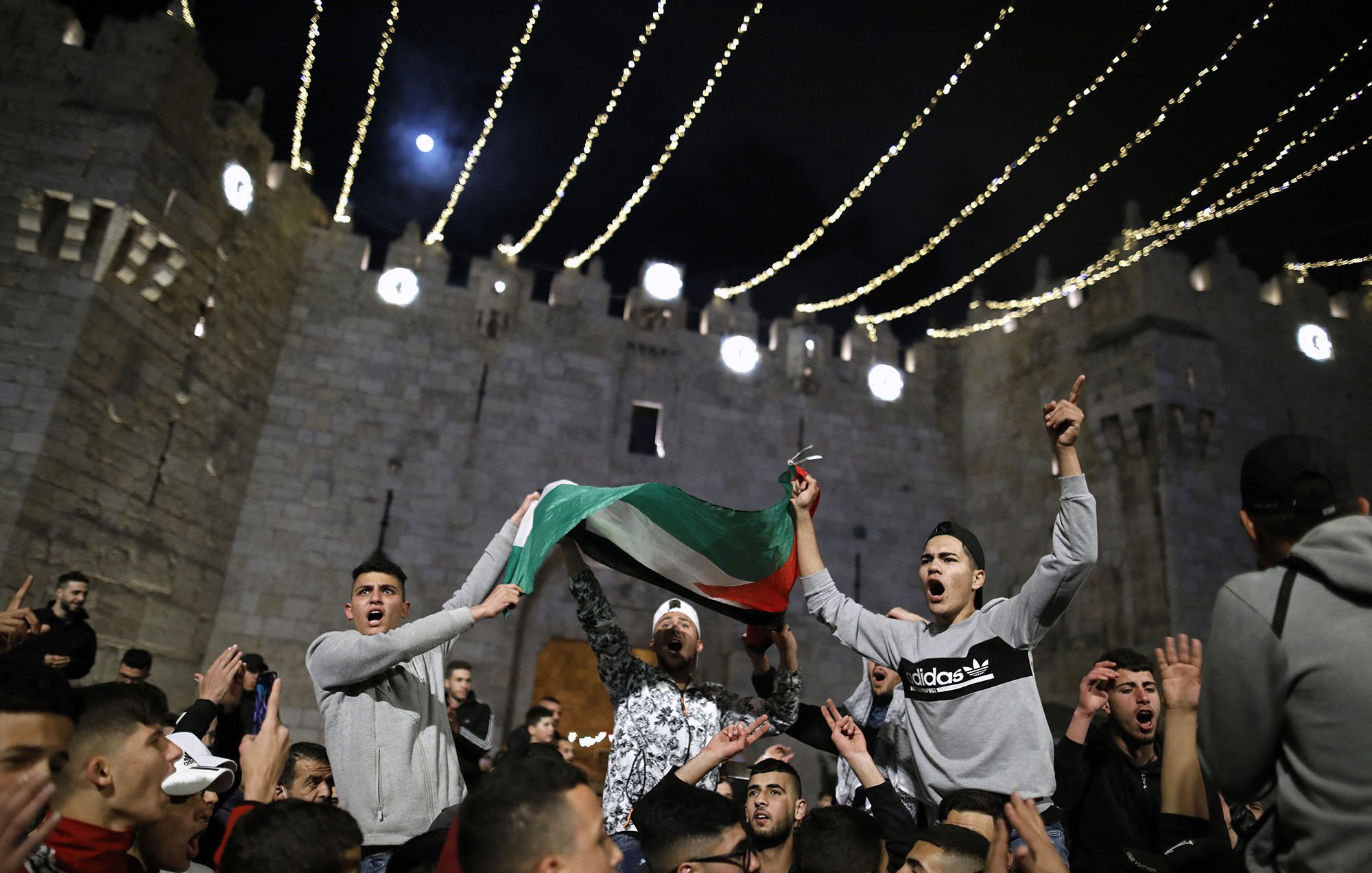 L'immagine mostra un gruppo di persone riunite in un contesto festivo, probabilmente in un evento pubblico. I partecipanti stanno sventolando una bandiera, che sembra essere quella palestinese, e sono visibilmente entusiasti. Sullo sfondo, si può notare una struttura medievale, che suggerisce un'atmosfera di celebrazione. Le luci appese creano un'atmosfera suggestiva e notturna, mentre i volti delle persone esprimono gioia e partecipazione.