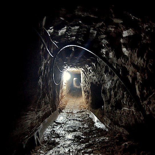 L'image montre un tunnel sombre creusé dans la terre. Les parois de la cavité sont en pierre, et le sol est inégal, probablement en terre battue. Une lumière blanche éclaire partiellement le tunnel, révélant son intérieur. Le fond du tunnel semble s'étendre à l'infini, créant une ambiance mystérieuse. Des câbles électriques sont visibles à certains endroits, indiquant qu'il pourrait y avoir des installations techniques à l'intérieur.