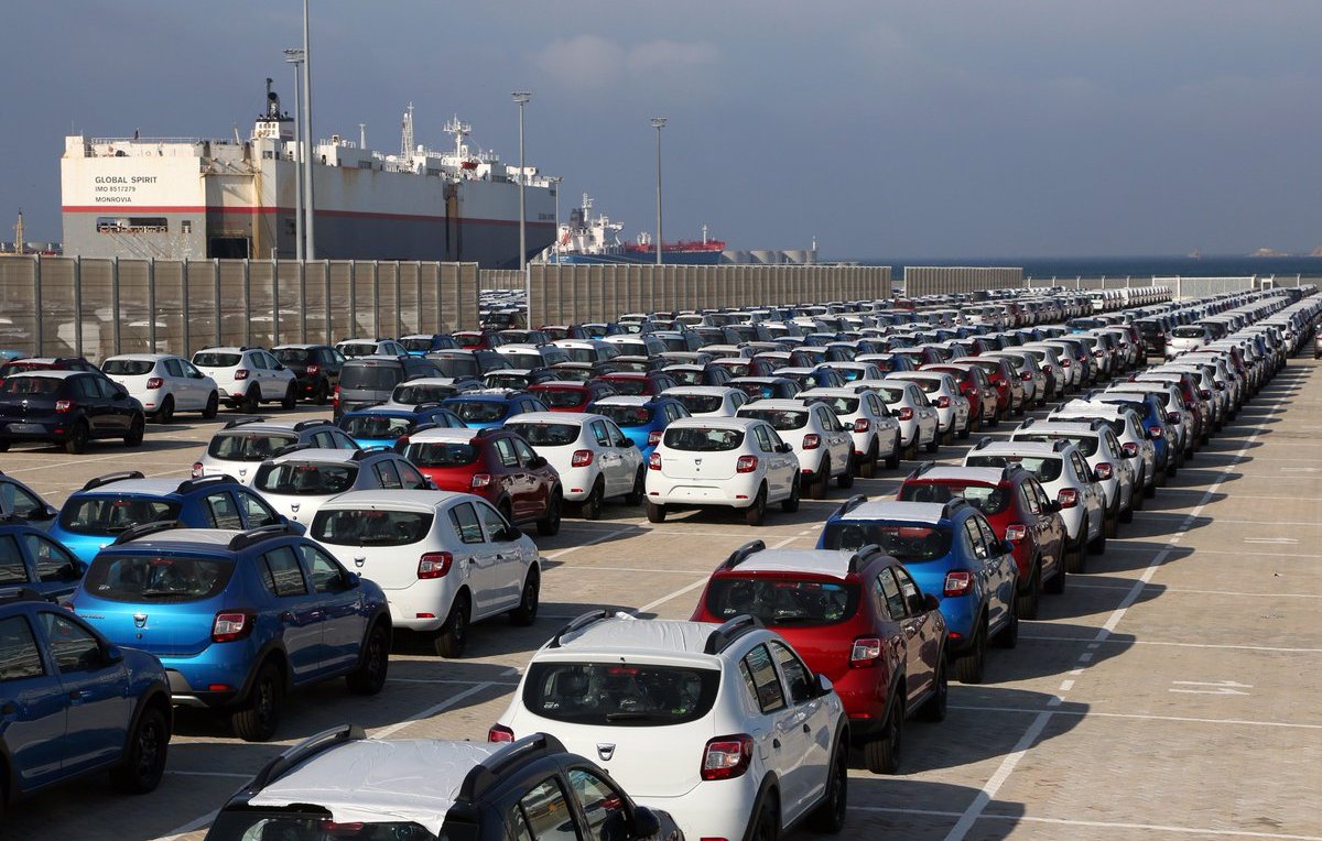 L'image montre un grand parking de voitures, organisé en rangées bien alignées. On aperçoit plusieurs types de voitures de différentes couleurs, majoritairement en blanc, rouge et bleu. En arrière-plan, on peut voir des bateaux de marchandises amarrés dans un port, sous un ciel nuageux. Cette scène semble se dérouler dans une zone portuaire, où les véhicules sont probablement en attente d'expédition ou de distribution.