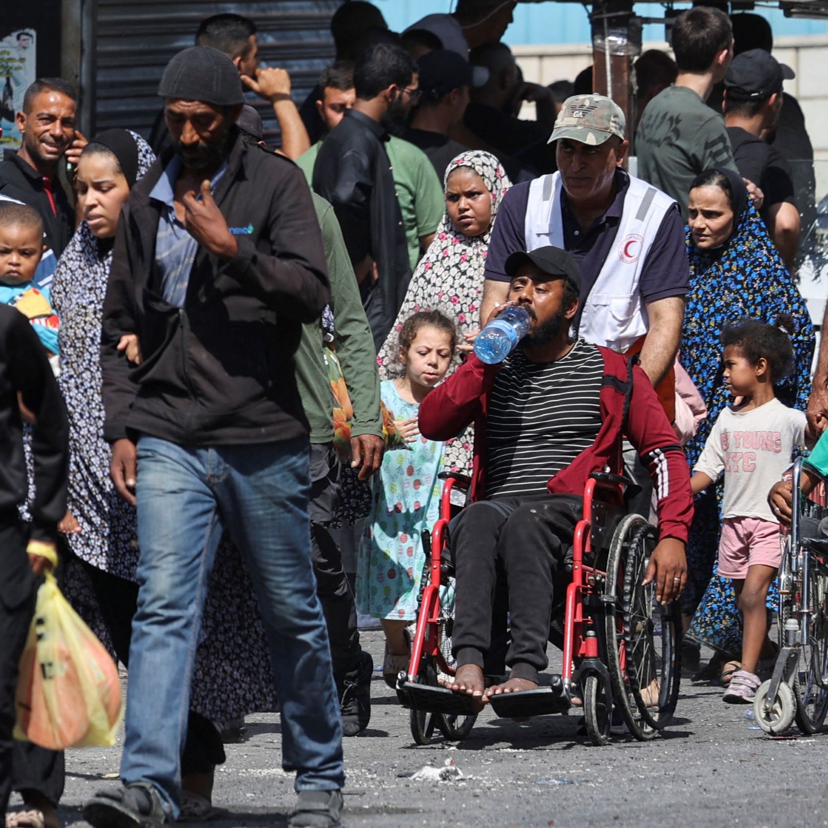 تظهر الصورة مجموعة من الأشخاص يسيرون في منطقة مزدحمة. يوجد رجال ونساء وأطفال، وبعضهم يظهر في كراسي متحركة. يتواجد في الخلفية بعض المحلات والبائعين. يبدون مشغولين أو في طريقهم إلى مكان ما، مما يعكس حركة الحياة اليومية في هذا المكان.