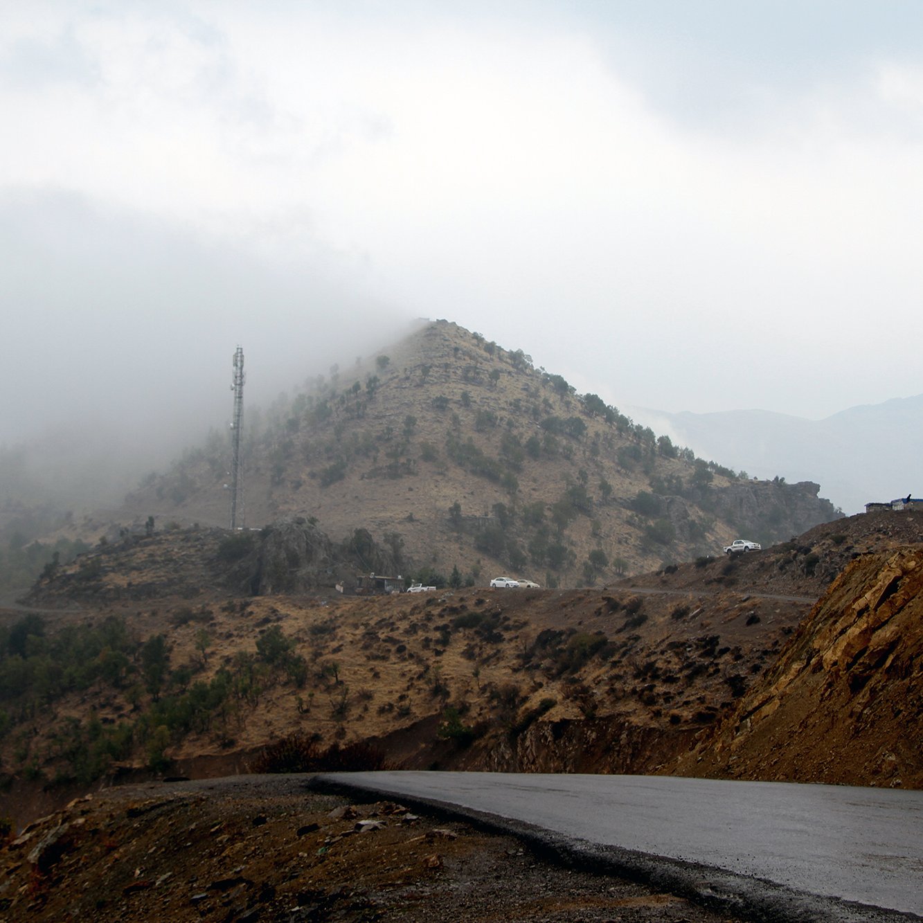 The image depicts a winding road leading through a mountainous landscape. In the background, a hill covered with low vegetation is partially obscured by mist or fog, creating a mysterious atmosphere. Atop the hill, there is a communication tower, and some vehicles are visible near the summit. The foreground features a dark, asphalt road that curves, suggesting a remote and rugged terrain. The overall scene conveys a sense of tranquility and solitude in nature.