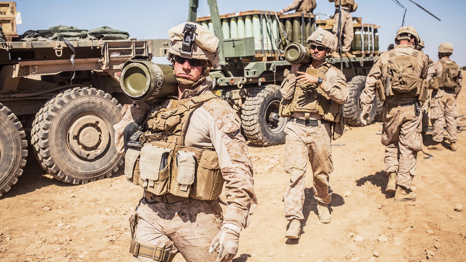 L'image montre des soldats en tenue militaire, probablement en opération. Certains d'entre eux portent des gilets tactiques et des casques, tandis qu'un soldat en avant tient un obus de mortier. En arrière-plan, on aperçoit un véhicule militaire avec une remorque et d'autres soldats s'affairant. Le cadre semble désertique, avec un sol sablonneux et un ciel dégagé. On ressent un climat de préparation et d'activité militaire.