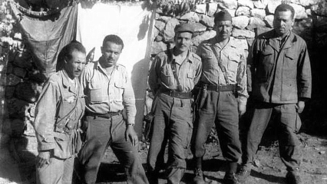 The image shows a group of five soldiers standing together in a rural setting. They are wearing military uniforms and appear to be posing for the photo. The background consists of a stone wall and a makeshift tarp or cloth, suggesting a temporary encampment. The soldiers have serious expressions, reflecting the context of their environment. Overall, the image conveys a sense of camaraderie among them in a military setting.