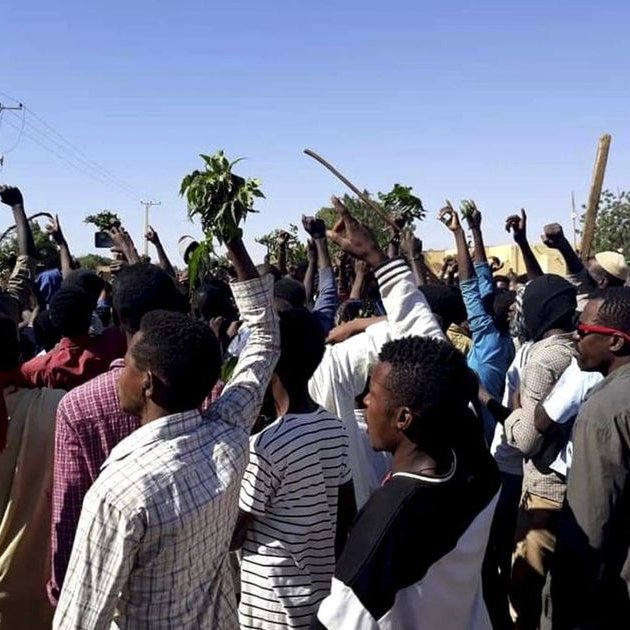 L'image montre un grand groupe de personnes rassemblées à l'extérieur, levez les bras en signe de protestation ou de soutien, tenant probablement des branches ou des plantes. Le paysage semble être rural, avec quelques bâtiments en arrière-plan et un ciel dégagé. L'ambiance semble dynamique et énergique, reflétant une mobilisation collective.