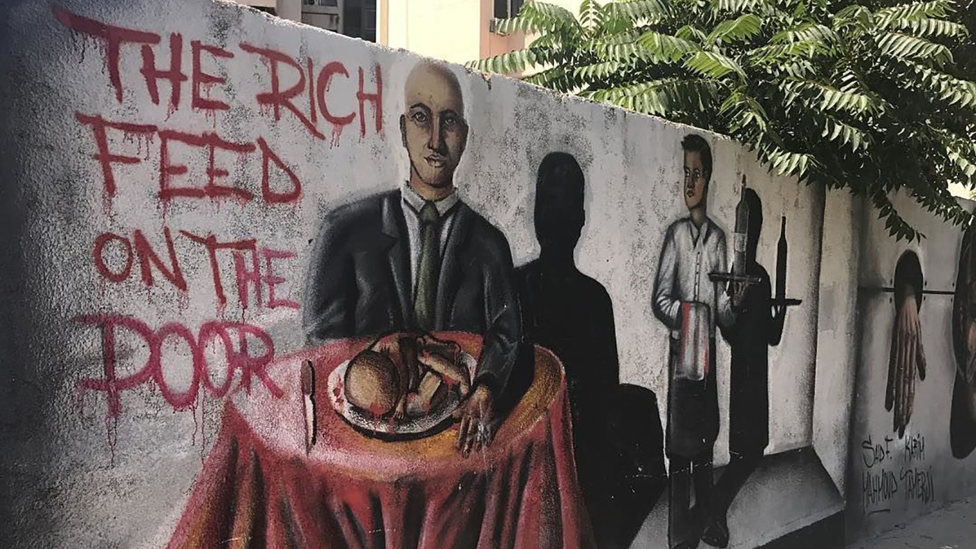 The image features a mural on a wall depicting two main figures. On the left, a man in a suit sits at a table covered with a red tablecloth, holding a loaf of bread and looking directly at the viewer. To the right, another figure dressed as a waiter stands with a tray, while dark shadows cast behind them suggest more figures or a hidden narrative. The phrase "THE RICH FEED ON THE POOR" is painted prominently nearby, conveying a theme of social inequality and critique of class disparity. The background includes urban elements like buildings and greenery, adding to the setting of the mural.