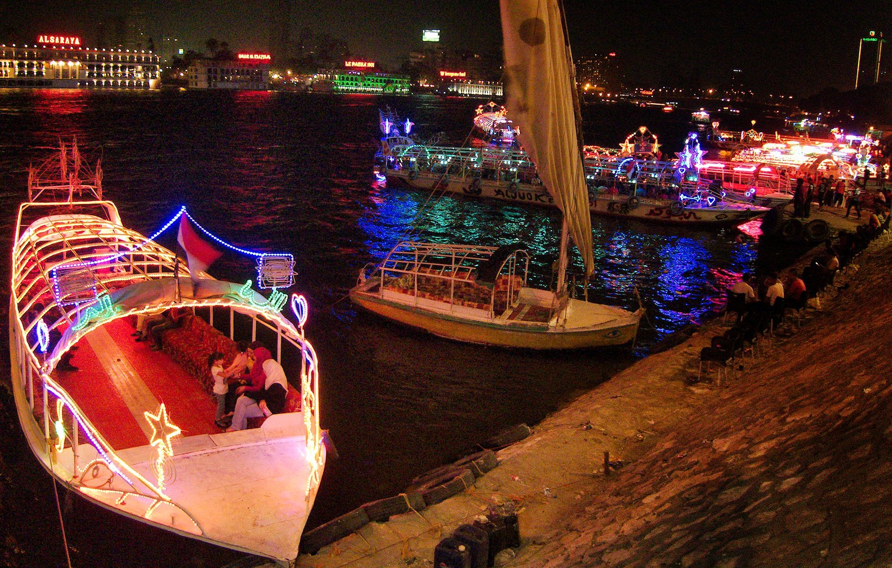L'immagine mostra una scena notturna su un fiume, con diverse barche decorate con luci colorate. Le imbarcazioni si trovano in una zona animata, e il riflesso delle luci sull'acqua crea un'atmosfera vivace e festosa. Sullo sfondo, si possono vedere edifici illuminati, che contribuiscono ulteriormente all'ambiente festivo della scena. Alcune persone sono sedute lungo la riva, godendosi la vista e l'atmosfera.