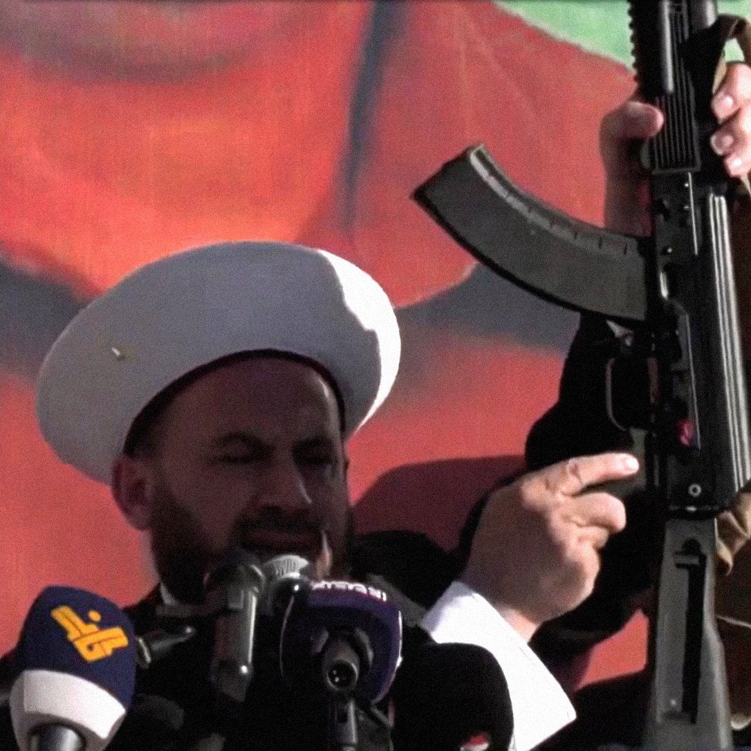 The image depicts two men, wearing white turbans and traditional garments, standing at a podium with microphones. One man is prominently holding an assault rifle above his head, while the other looks on. The background features a large, colorful mural or poster, possibly of a political figure or symbol. The scene suggests a demonstration or rally, conveying themes of power and assertiveness.