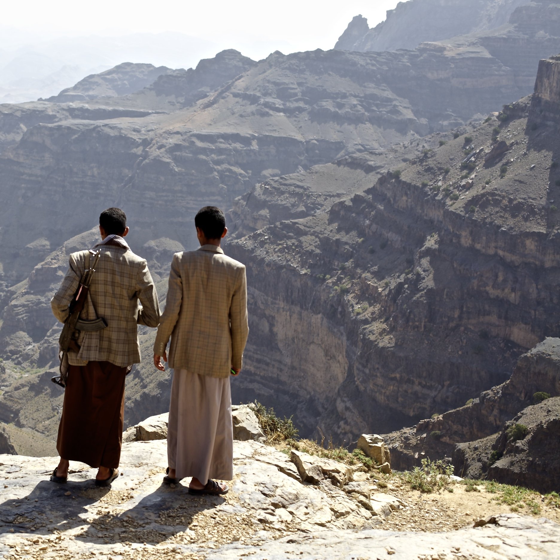 L'immagine mostra due uomini che si trovano su un bordo roccioso, ammirando un vasto paesaggio montano. Le loro spalle sono visibili mentre guardano verso il fondo di una valle profonda e frastagliata. Le montagne circostanti sono di diverse tonalità di grigio e marrone, con una vegetazione scarsa visibile in alcune zone. L'atmosfera appare serena e suggestiva, con un cielo chiaro sullo sfondo.