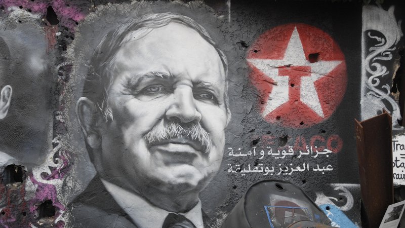 L'image montre un graffiti sur un mur, représentant un portrait en noir et blanc d'un homme avec une moustache et un regard déterminé. À côté du portrait, on peut voir une étoile rouge avec un symbole. Des inscriptions en arabe sont également visibles, accompagnées de motifs colorés sur le mur. L'ensemble donne une impression d'engagement politique, avec une ambiance urbaine et artistique.