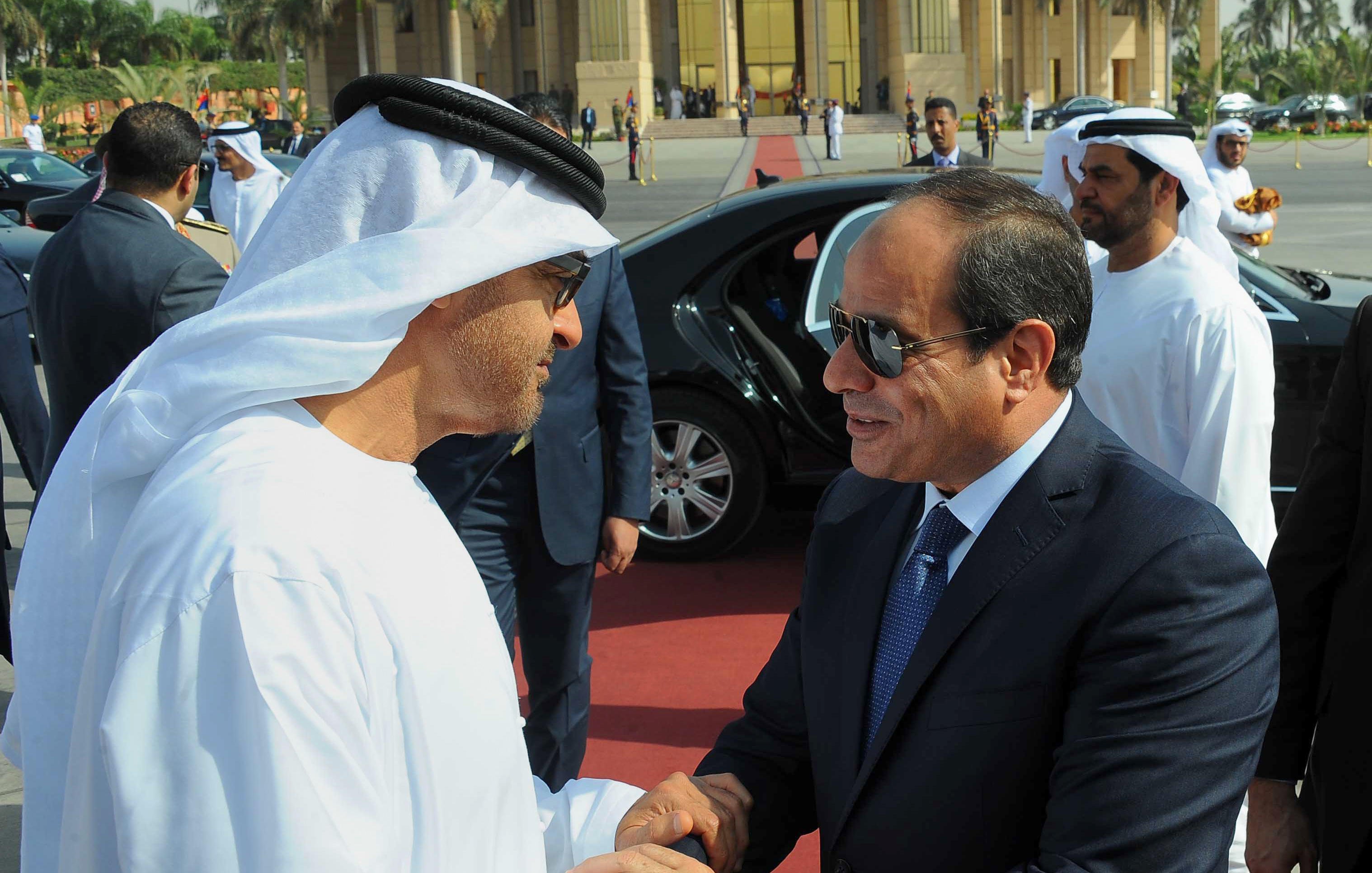 The image depicts two men in formal attire engaged in a handshake, suggesting a cordial greeting. One man is dressed in traditional Emirati clothing, including a white kandura and headscarf, while the other is wearing a dark suit and tie. They appear to be in an official setting, with a luxurious car and a group of well-dressed individuals in the background. The atmosphere indicates a diplomatic or state visit, emphasizing cooperation and respect between the two figures.