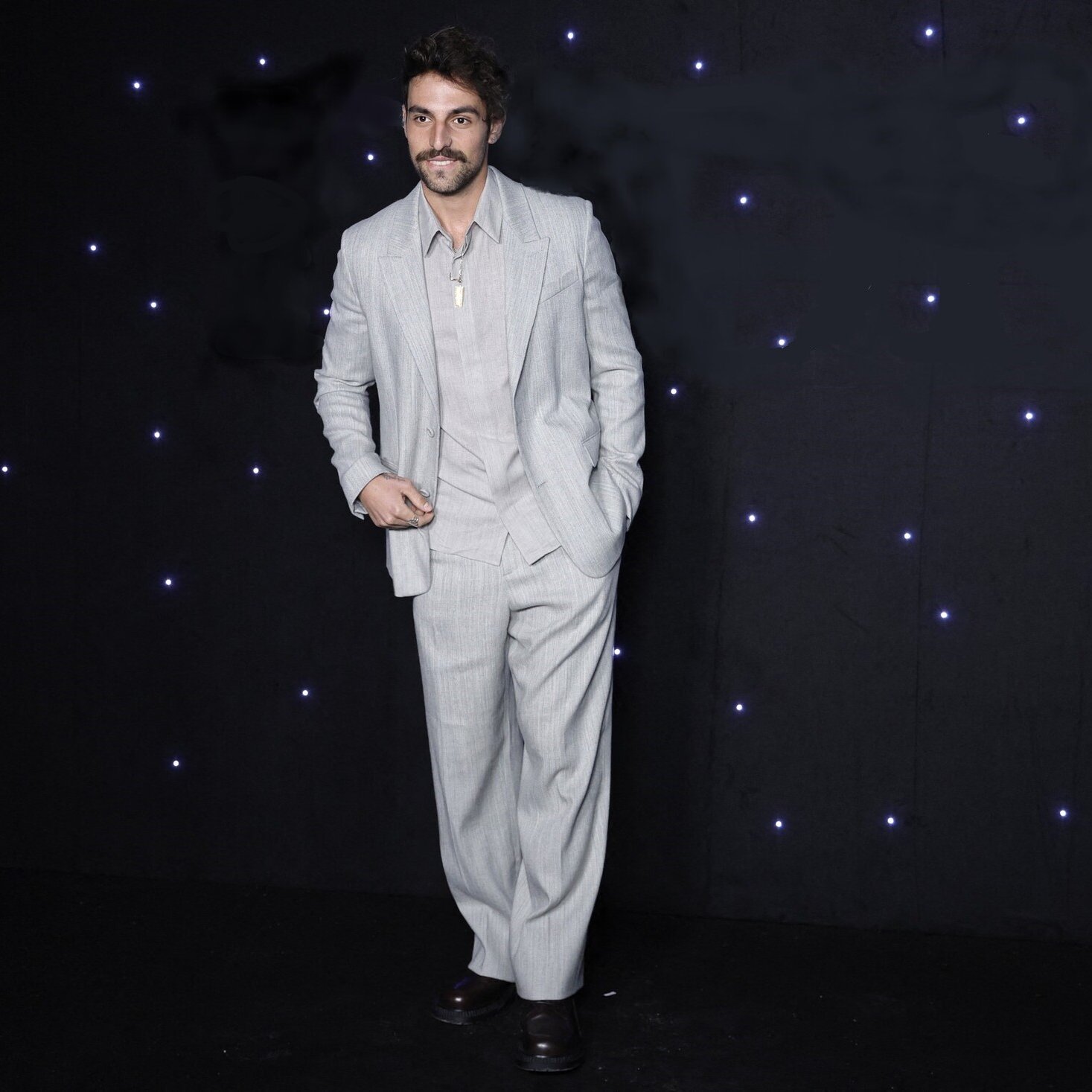 La imagen muestra a un hombre vestido con un traje claro de color gris. El traje tiene un corte elegante, con pantalones de tiro alto y una camisa debajo que parece ser de un tono similar. El hombre tiene una expresión sonriente y está posando de manera confiada, con una mano en el bolsillo del pantalón. El fondo es oscuro y está adornado con luces brillantes que aportan un toque festivo a la escena.