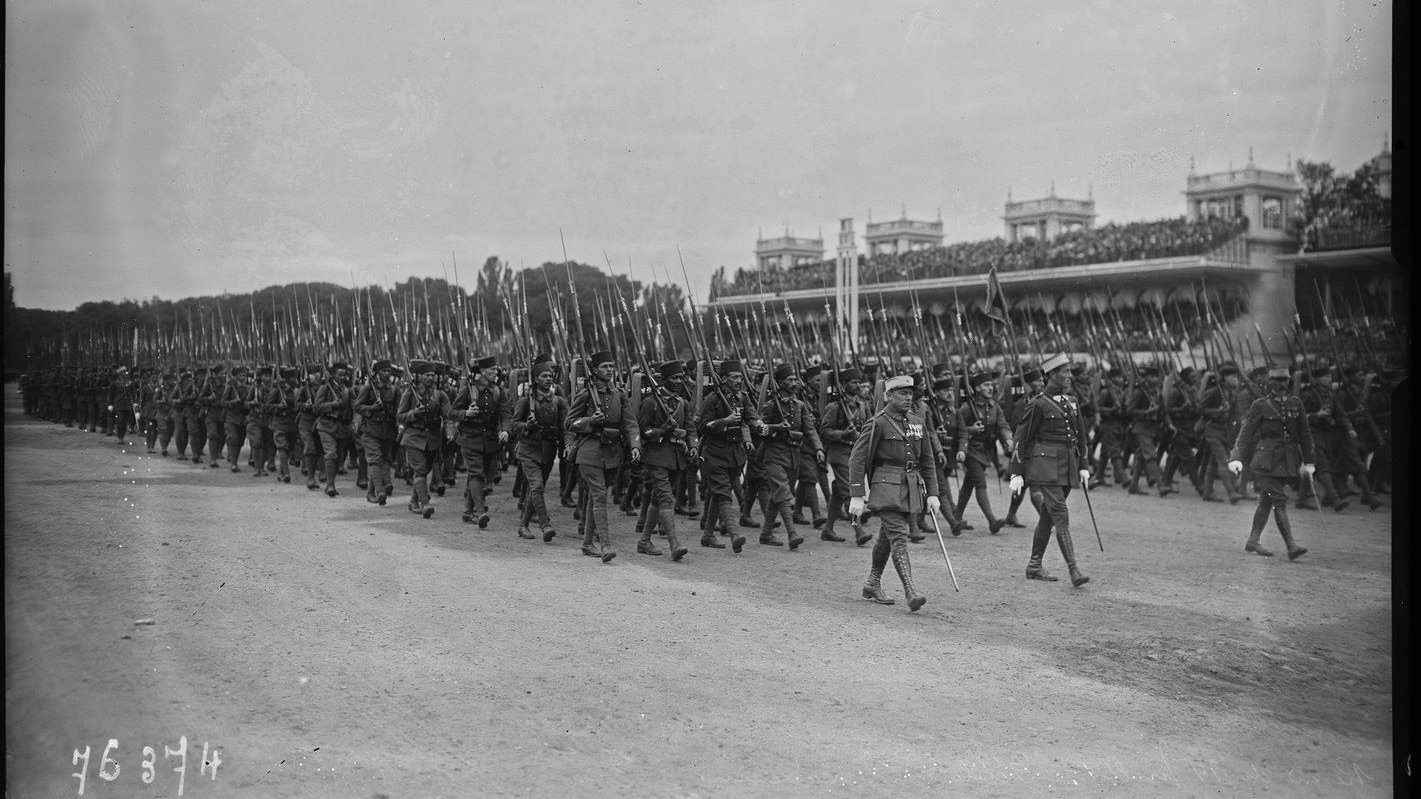 L'image montre un défilé militaire, avec de nombreux soldats marchant en formation. Ils portent des uniformes d'époque et tiennent des fusils à la main. En arrière-plan, on peut voir une tribune avec des spectateurs, probablement des dignitaires ou des spectateurs admirant le défilé. L'atmosphère semble solennelle et organisée, typique des événements militaires de ce genre. Le ciel est nuageux, ce qui donne une ambiance sobre à la scène.