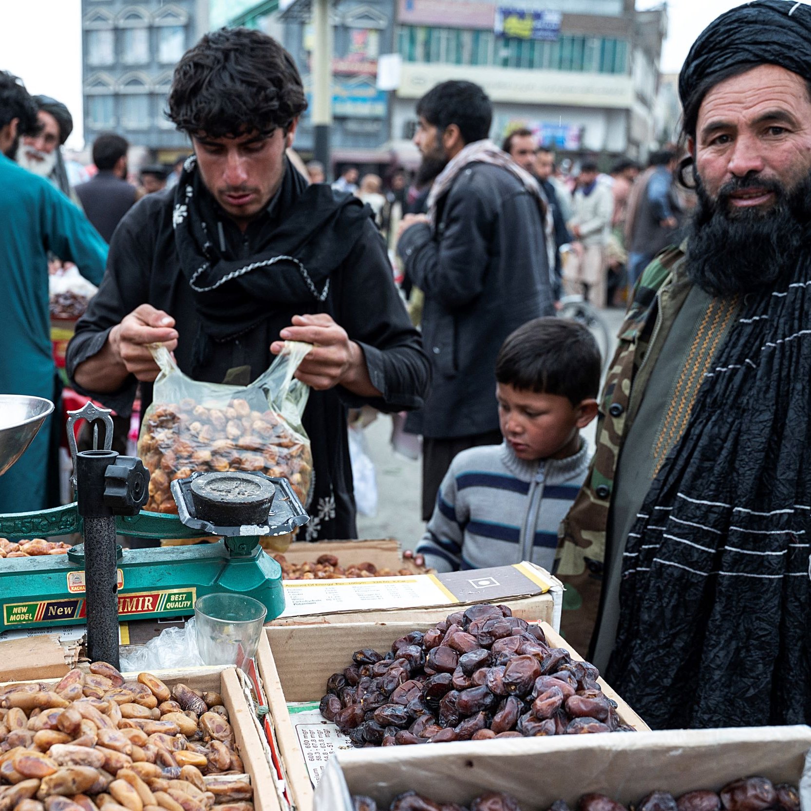 L'image montre un marché en plein air, probablement dans un pays asiatique. Au premier plan, on voit un homme portant un turban et une barbe, habillé d'une veste militaire. À côté de lui, un enfant observe. L'homme a l'air de s'occuper d'une balance et de dates, qui sont présentées dans des caisses en bois à ses côtés. En arrière-plan, on aperçoit d'autres personnes, ce qui donne une impression de foule animée et d'activité commerciale. L'environnement semble rural et traditionnel, avec des bâtiments flous au fond.