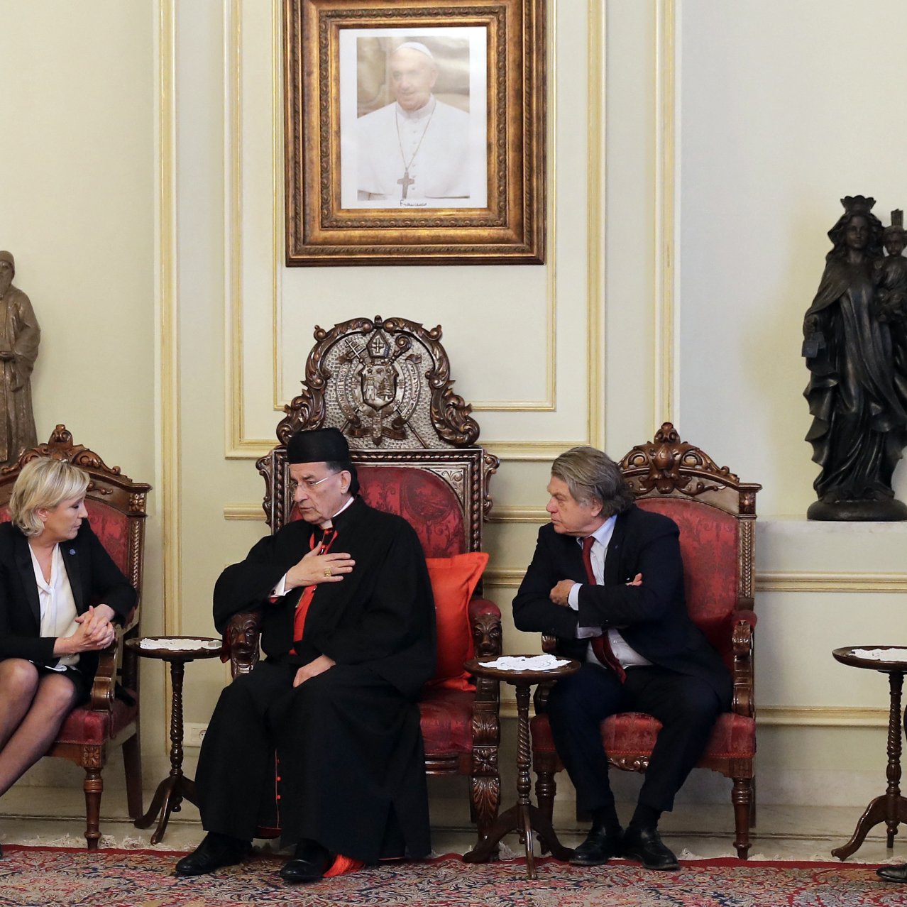 در این تصویر، یک جلسه‌ای در یک اتاق رسمی دیده می‌شود. در مرکز تصویر، یک مرد با لباس مذهبی نشسته است که ظاهراً یک مقام دینی است. او دستش را بر روی قلبش قرار داده و به دو نفر دیگر که در طرفین او نشسته‌اند، توجه می‌کند. زن سمت چپ تصویر گویی در حال گوش دادن به صحبت‌های اوست و مرد سمت راست تصویر نیز در حال بحث یا تبادل نظر به نظر می‌رسد. محیط اتاق تزئین شده و شامل یک تابلو در دیوار و مجسمه‌هایی در پس‌زمینه است. فرش زیر پای آنها نیز به فضای رسمی‌ و سنتی اتاق افزوده است.