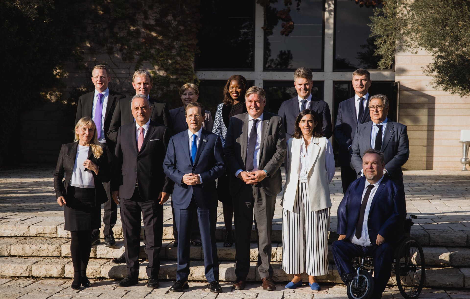 الصورة تظهر مجموعة من الأشخاص يقفون معًا في بيئة خارجية، ربما تكون حديقة أو منطقة أمامية لمبنى. يوجد في الخلفية مشهد طبيعي مع أشجار ونباتات. يرتدي الحضور ملابس رسمية وأنيقة، مما يشير إلى أن الصورة قد تم التقاطها في مناسبة رسمية أو مؤتمر. تتراوح تعبيرات الوجوه بين الابتسامات والجدية، مما يعكس جوًا من التعاون والتواصل.