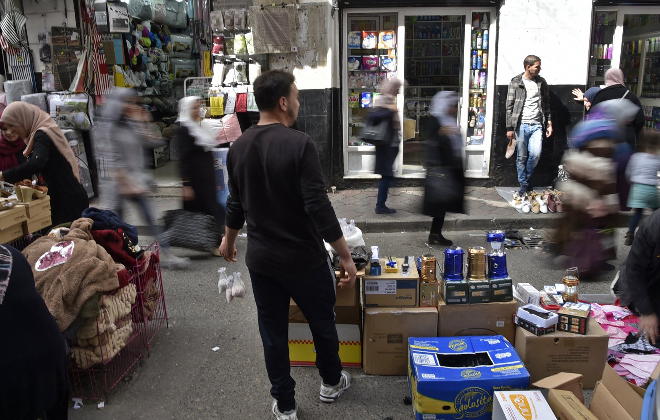 L'image montre une scène animée d'un marché urbain. On peut voir plusieurs personnes se déplacer rapidement, créant un effet de flou. Au premier plan, un homme se tient de dos, tenant des objets. Des étals sont visibles, avec des boîtes et des marchandises disposées sur des chariots. En arrière-plan, des magasins sont ouverts et on aperçoit des clients. L'atmosphère semble vivante et dynamique, typique d'un marché fréquenté.