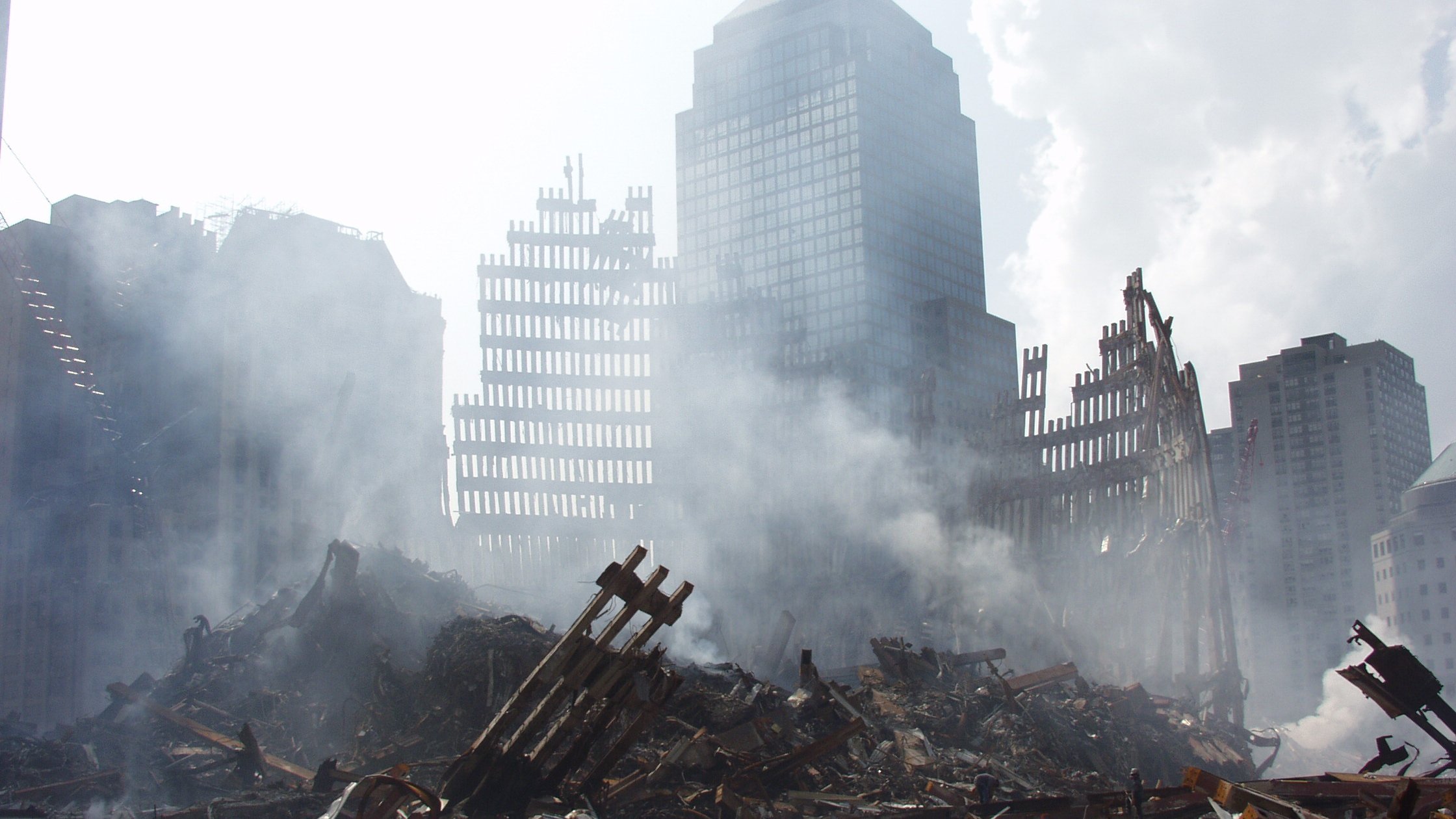 تصویر مربوط به یک صحنه خرابی و ویرانی است. در پس‌زمینه، ساختمان‌های بلند و ناتمام دیده می‌شوند که برخی از آنها به شدت آسیب‌دیده‌اند. در جلوی تصویر، خرابه‌هایی از سازه‌ها و تیرک‌های فلزی با دودی که از میان آنها بلند می‌شود، مشاهده می‌شود. حالت کلی تصویر نشان‌دهنده یک حادثه ویرانگر است و فضای ملتهب و غم‌انگیزی را ایجاد می‌کند.