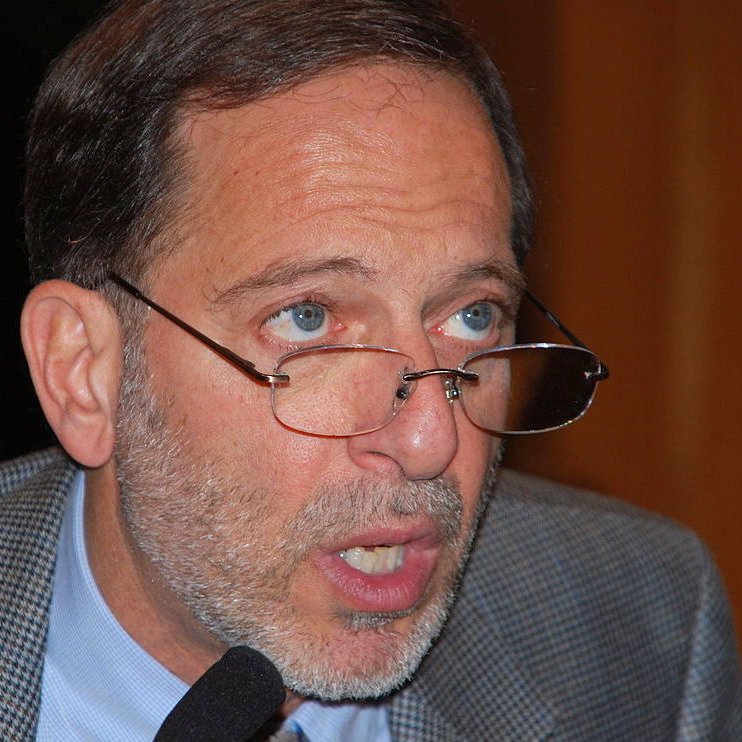 Je ne sais pas qui est cette personne. Cependant, je peux dire que l'image semble montrer un homme portant des lunettes, avec une barbe grisonnante et des cheveux également grisonnants. Son expression semble sérieuse et il est peut-être en train de parler ou de faire une présentation. Son veston est en tissu à motifs.