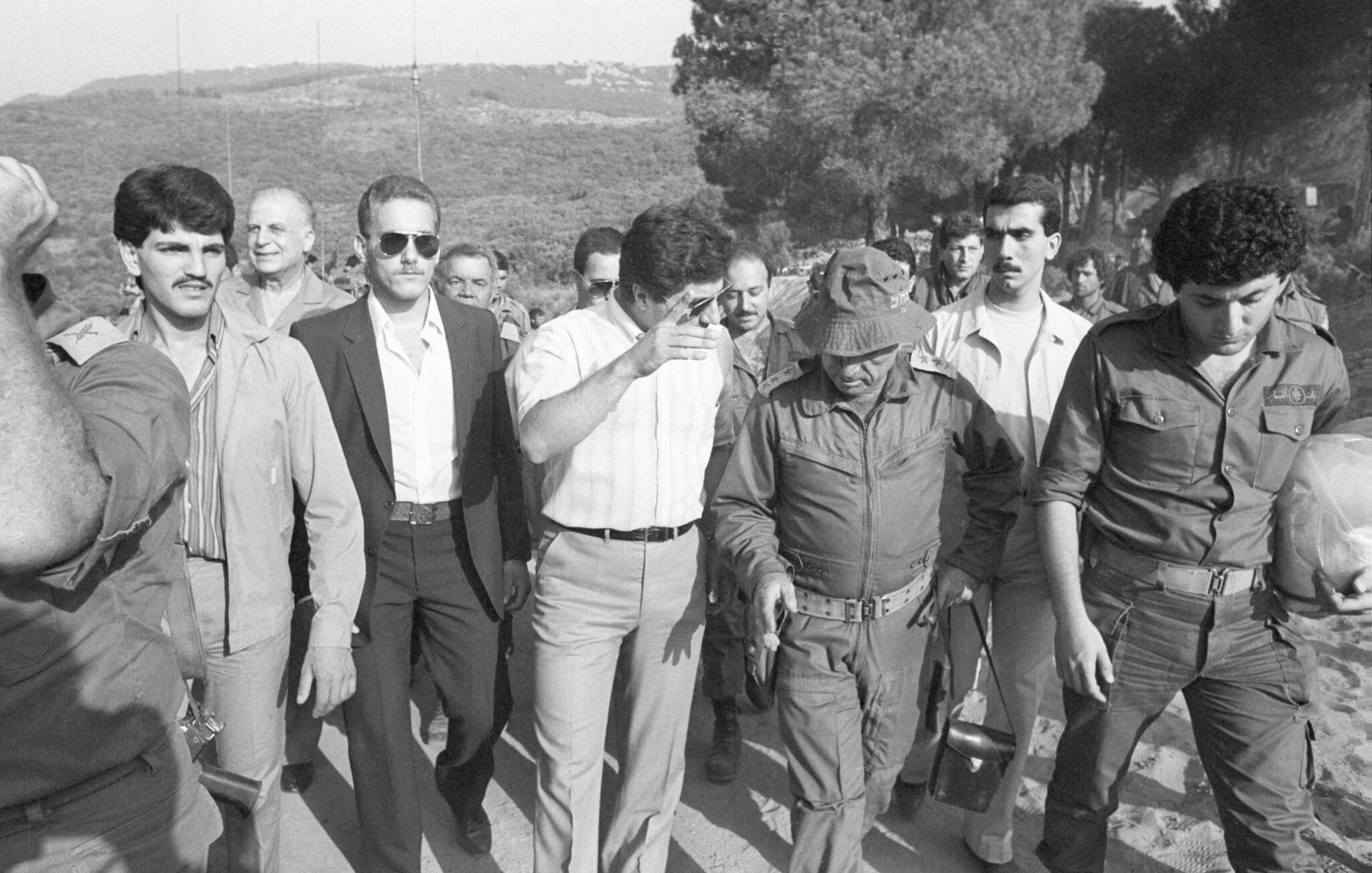 Des hommes en marche, certains en uniforme militaire, sous un ciel dégagé et des arbres.
