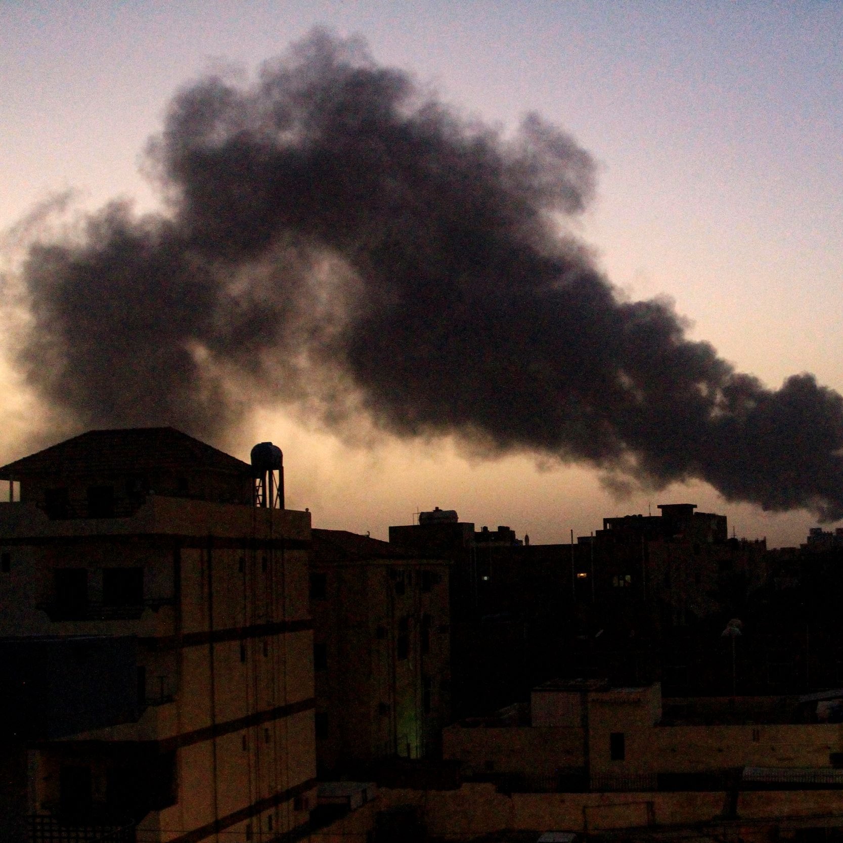 L'immagine mostra un paesaggio urbano al crepuscolo, con un cielo che tende al blu scuro. In lontananza si solleva una colonna di fumo nero, probabilmente proveniente da un incendio o una fonte di combustione. Gli edifici in primo piano sono silhouette scure contro il cielo, e si possono scorgere alcune luci artificiali che brillano tra le strutture. La vegetazione, come le palme, è visibile sul lato destro, aggiungendo un tocco di colore al contesto.